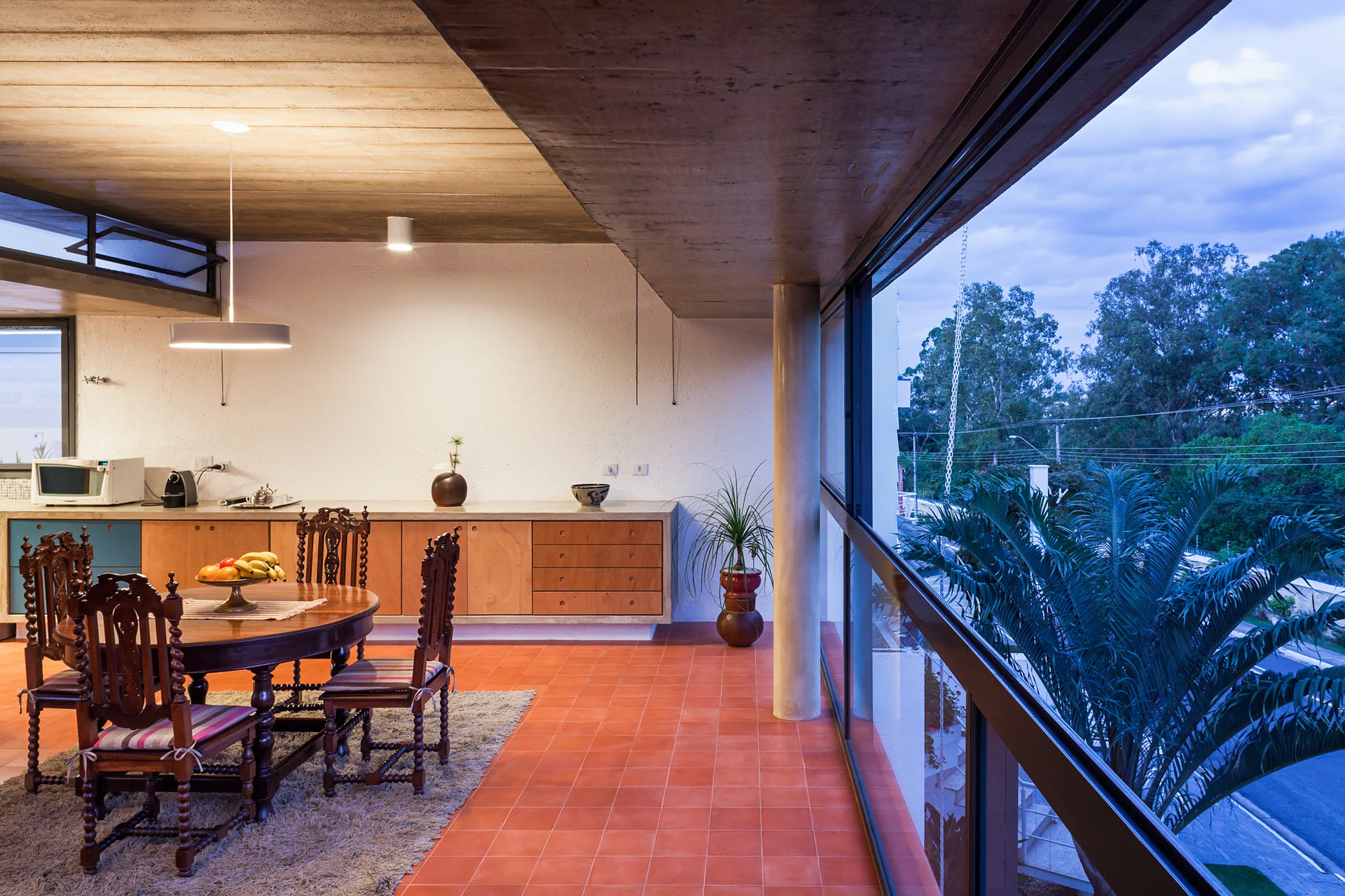 An open dining room features an ornate wooden table and chairs on a shag rug, set beneath an exposed concrete ceiling with terracotta tile floors.