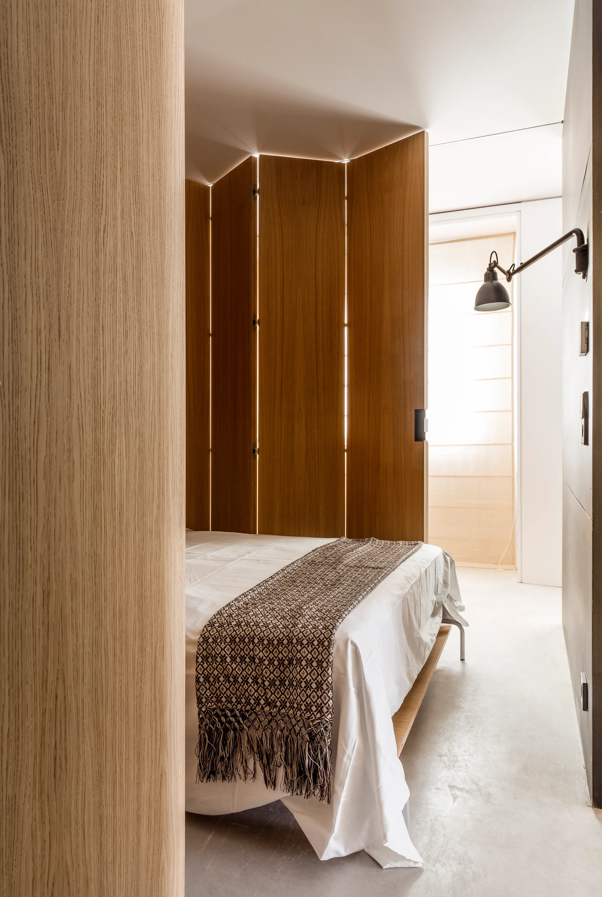 A minimalist bedroom with a white bed, a patterned brown throw, and a tall wooden folding screen serving as a partition.