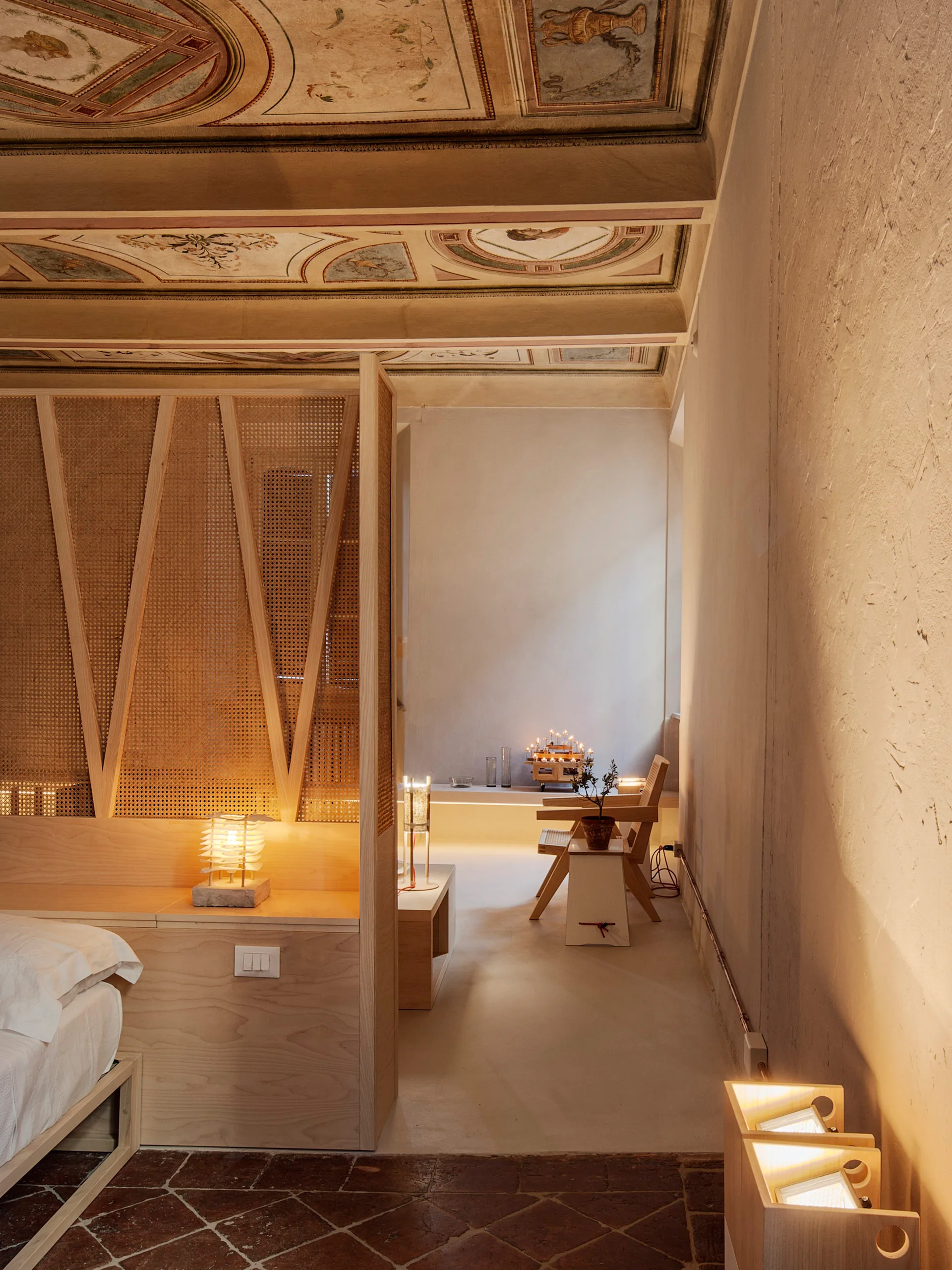 A modern bedroom features a light wood and cane partition, minimalist furniture, and terracotta floors beneath a historic painted fresco ceiling.