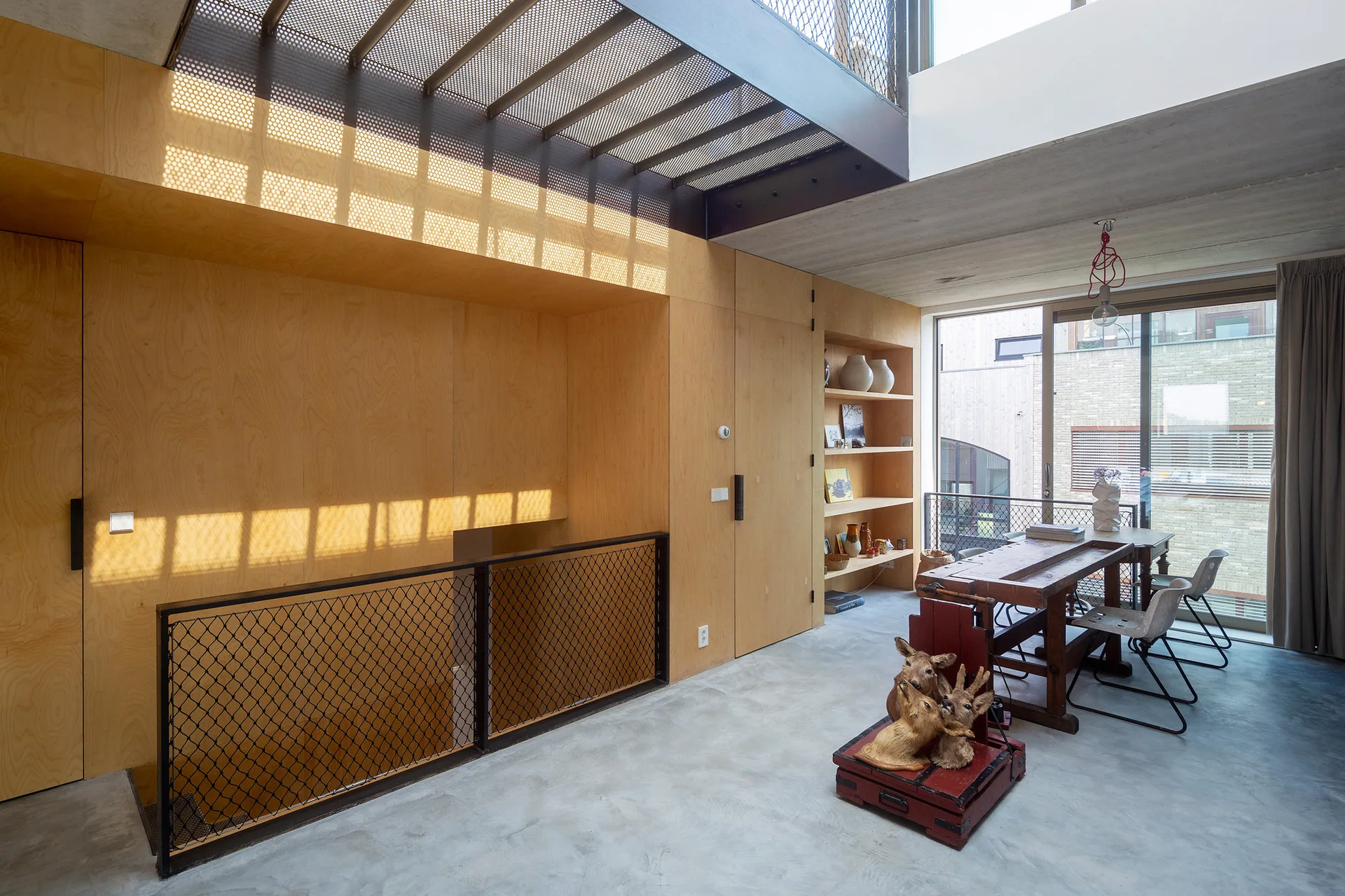 A modern loft dining area featuring a rustic wooden table, concrete flooring, built-in plywood shelving, and a metal staircase railing.