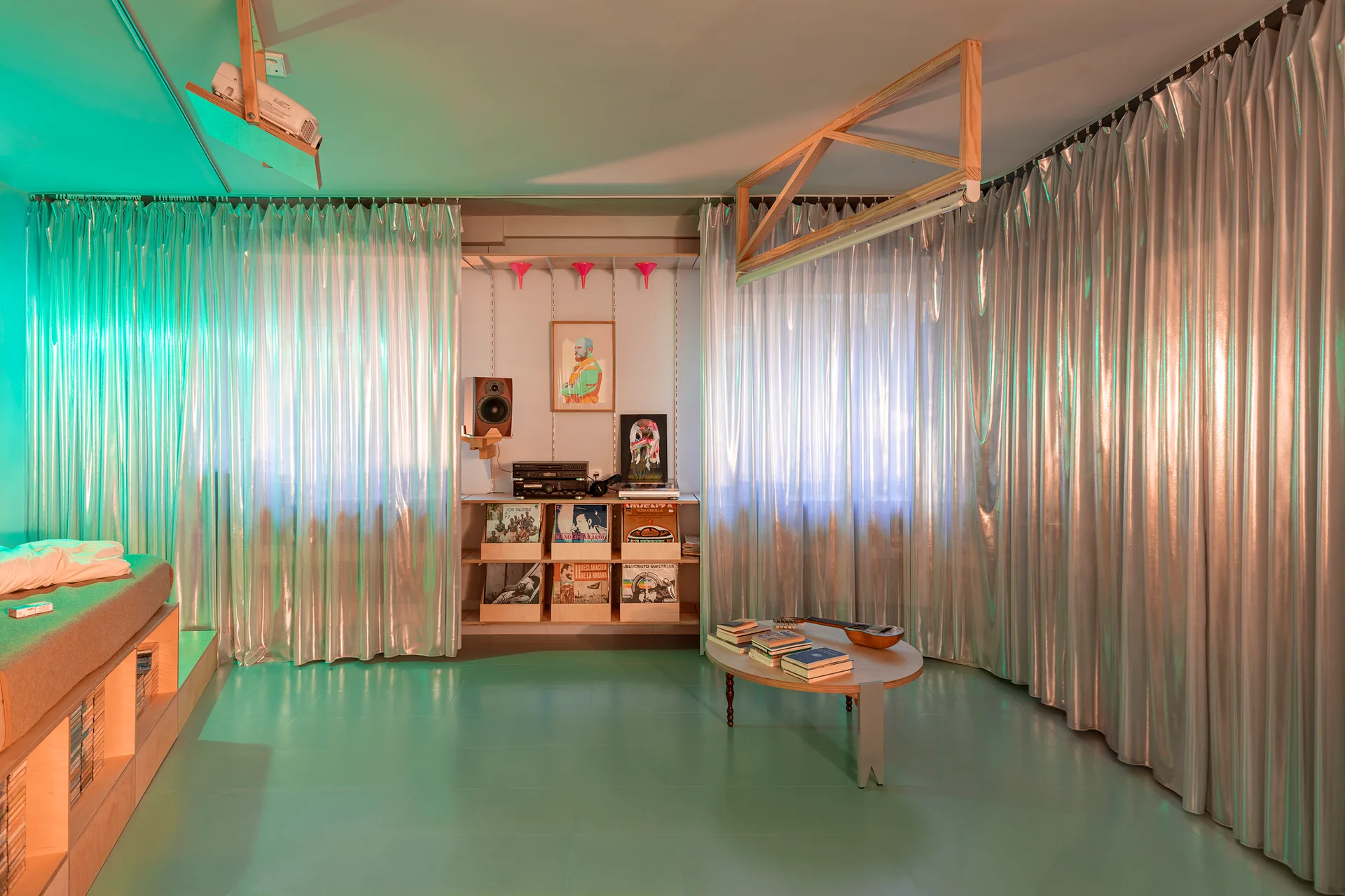 A media room features a green floor, custom plywood platform bed, low round wooden coffee table, and floor-to-ceiling metallic silver curtains.
