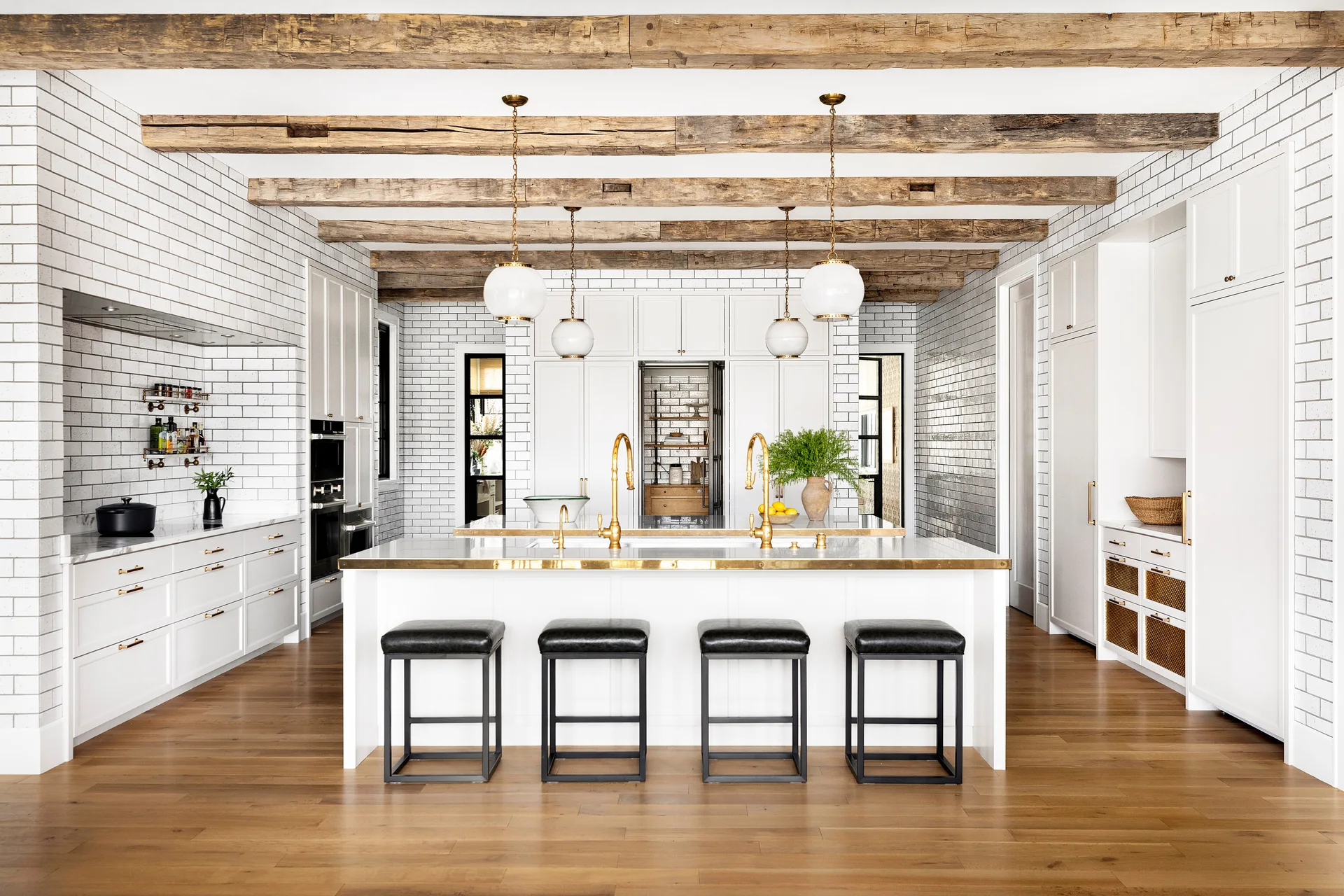 A bright kitchen featuring exposed wood ceiling beams, white subway tile walls, a white central island with brass trim, and black bar stools.