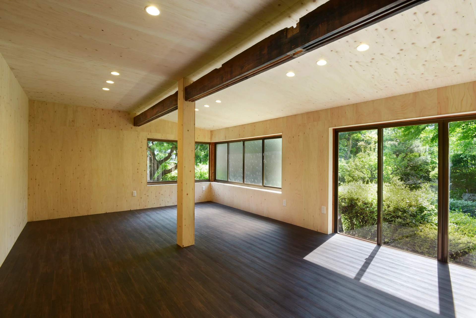 An empty room features light plywood walls and ceiling, a dark wood floor, a central support post, and glass doors looking onto a lush green garden.