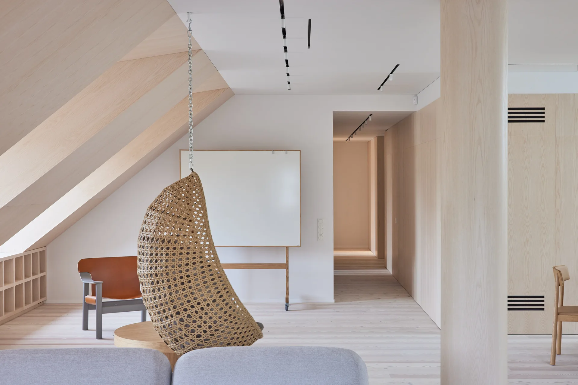 A minimalist room with a light wood sloped ceiling, wood paneling, a hanging woven chair, and a grey sofa in the foreground.