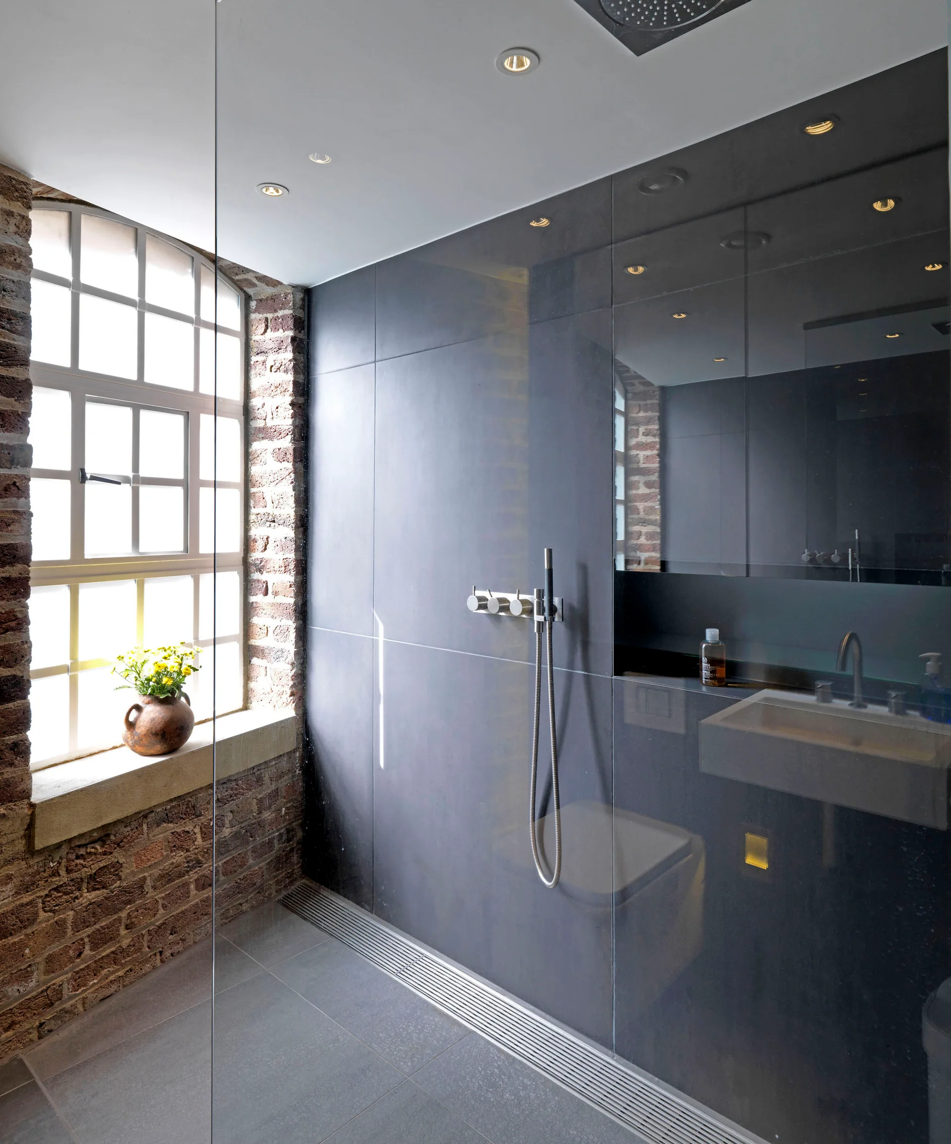 A modern industrial bathroom features a dark tiled shower enclosure, an exposed brick wall with an arched window, and a glass partition.