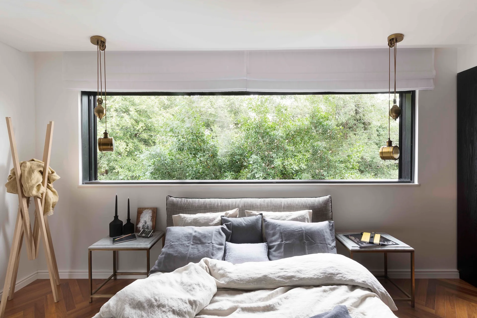 A bedroom with grey bedding under a wide horizontal window overlooking trees, flanked by brass pulley pendant lights and marble nightstands.