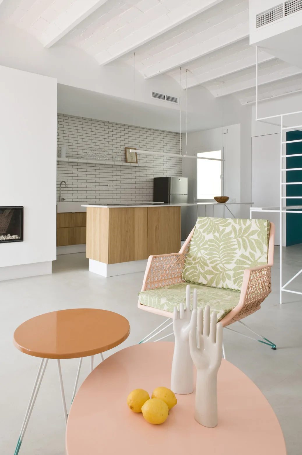 A bright open-plan space features a pink coffee table, a green patterned armchair, exposed white ceiling beams, and a kitchen with a wood island.
