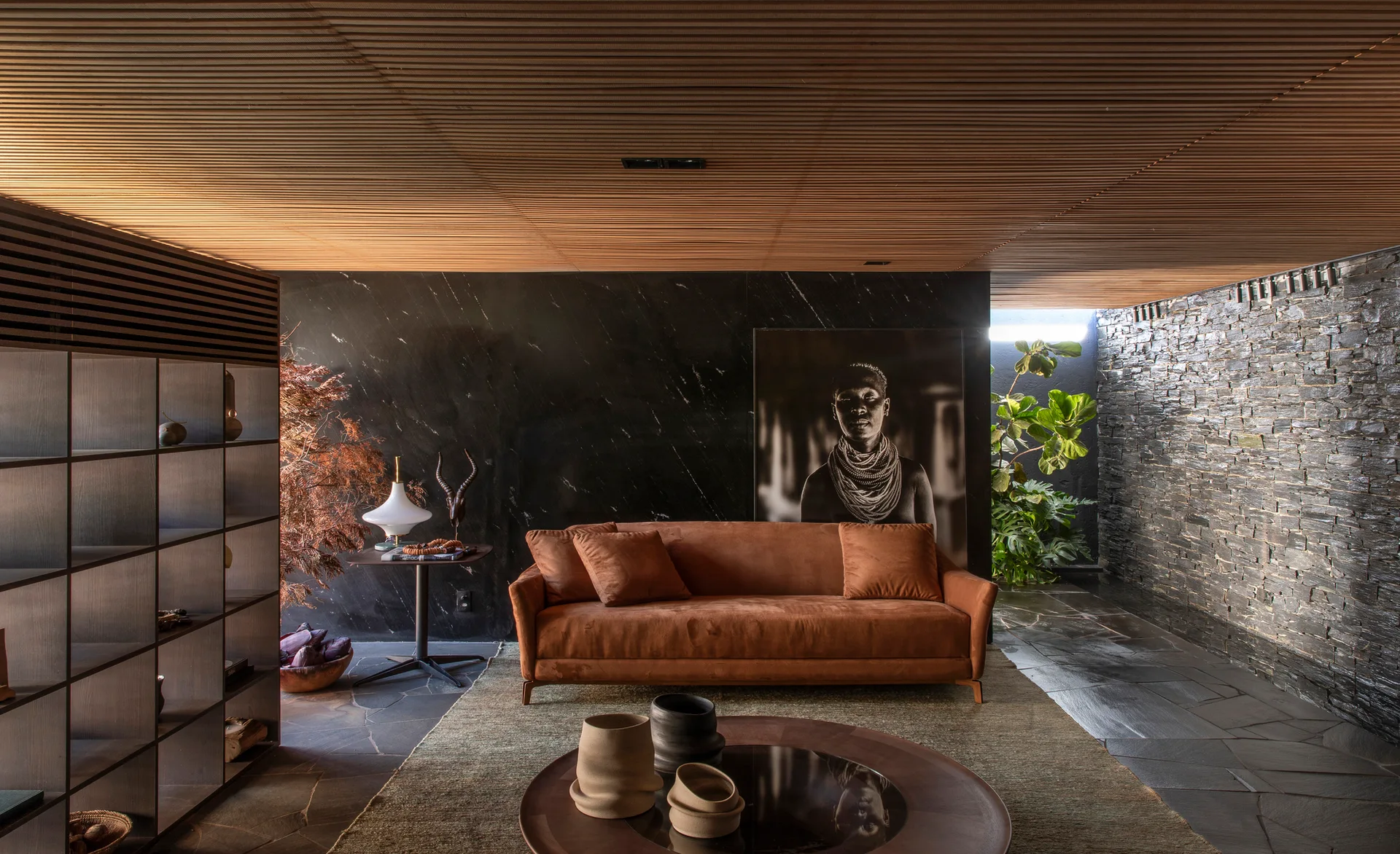 A modern living space features a rust-colored sofa against a black marble wall under a slatted wood ceiling, with a slate-tiled floor.