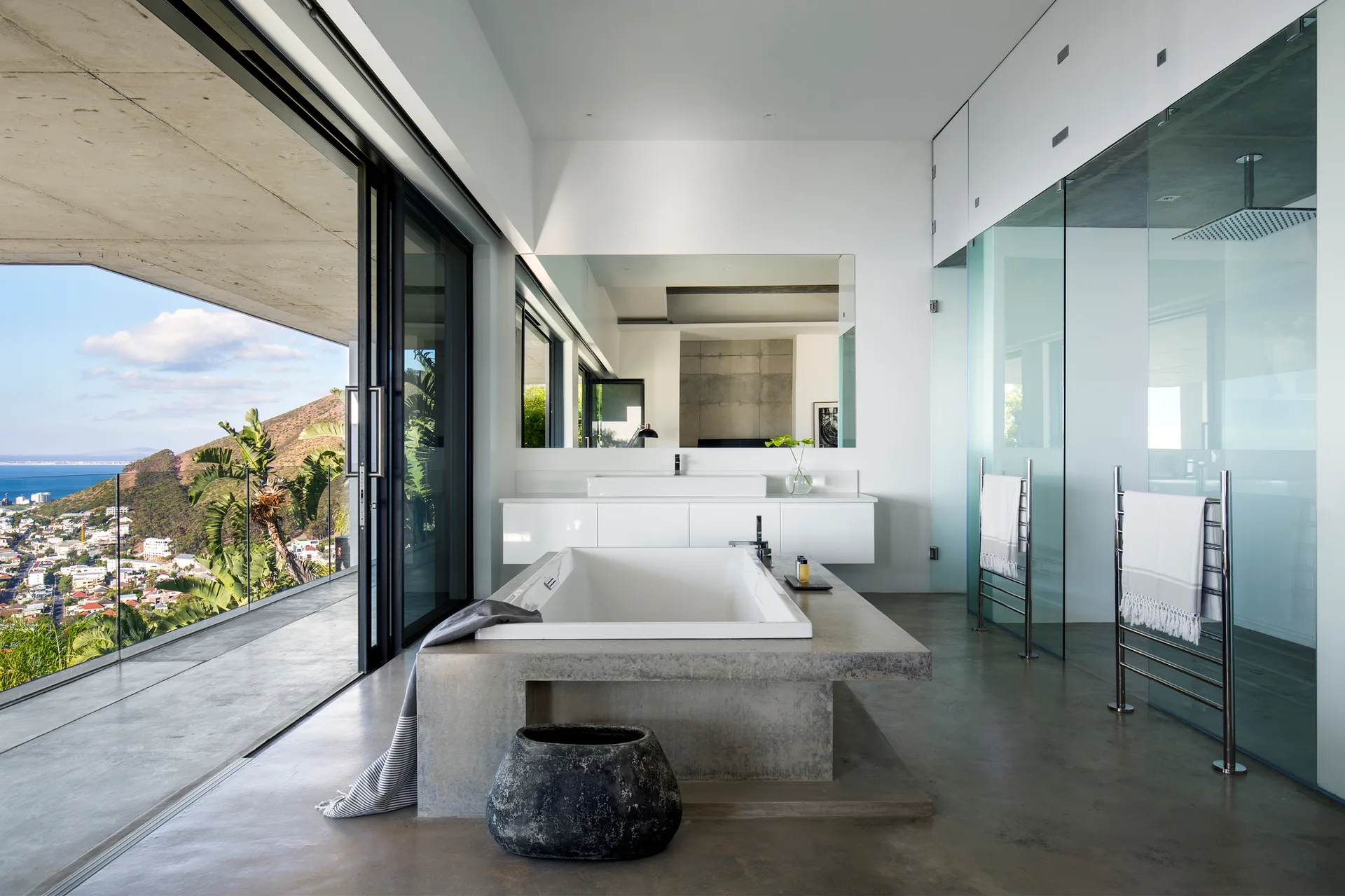 A modern bathroom with a central concrete-framed bathtub, a white floating vanity, and large glass sliding doors opening to an outdoor balcony.