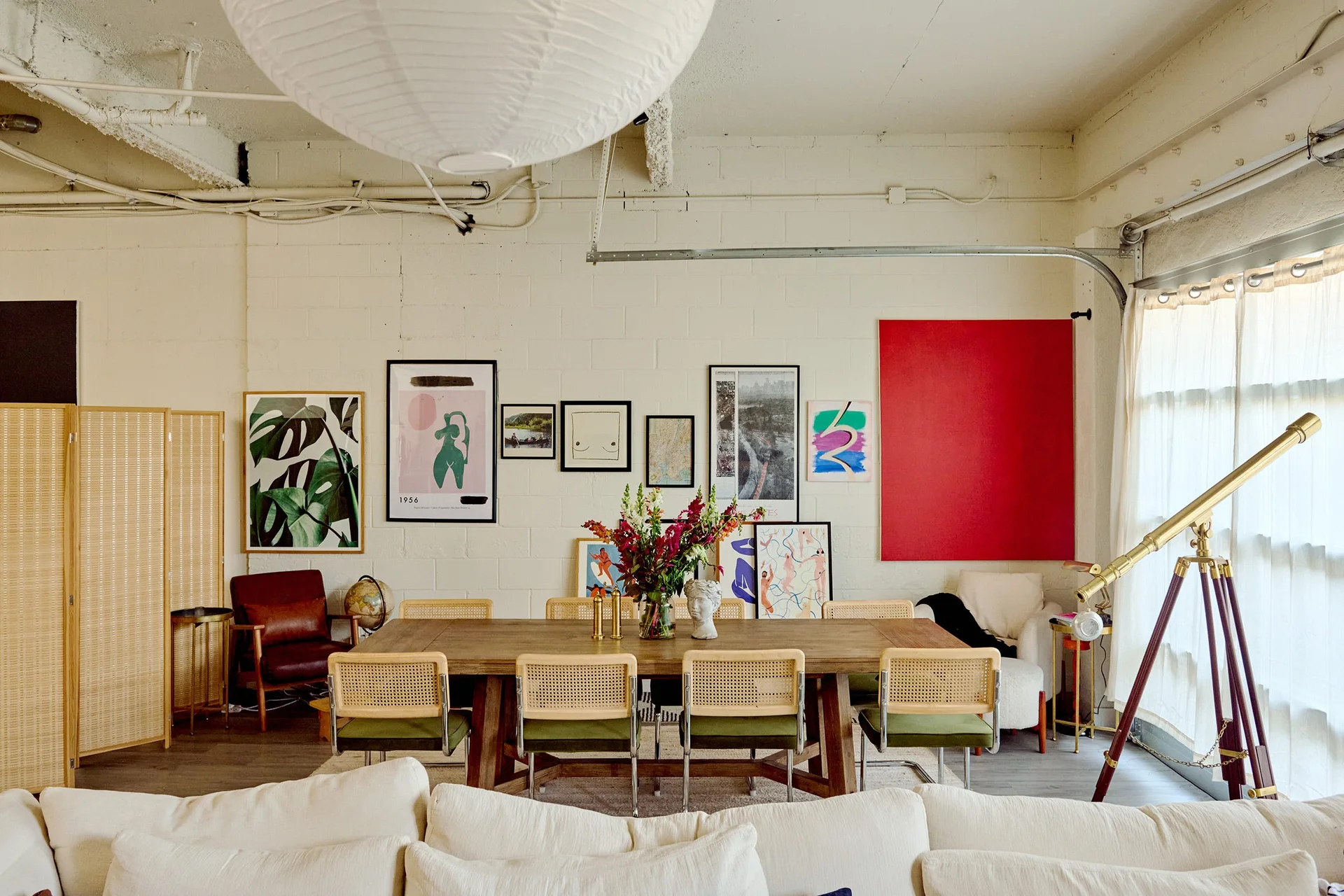 An eclectic dining room features a wooden table, woven cane chairs with green cushions, and a large gallery wall set against painted white brick.