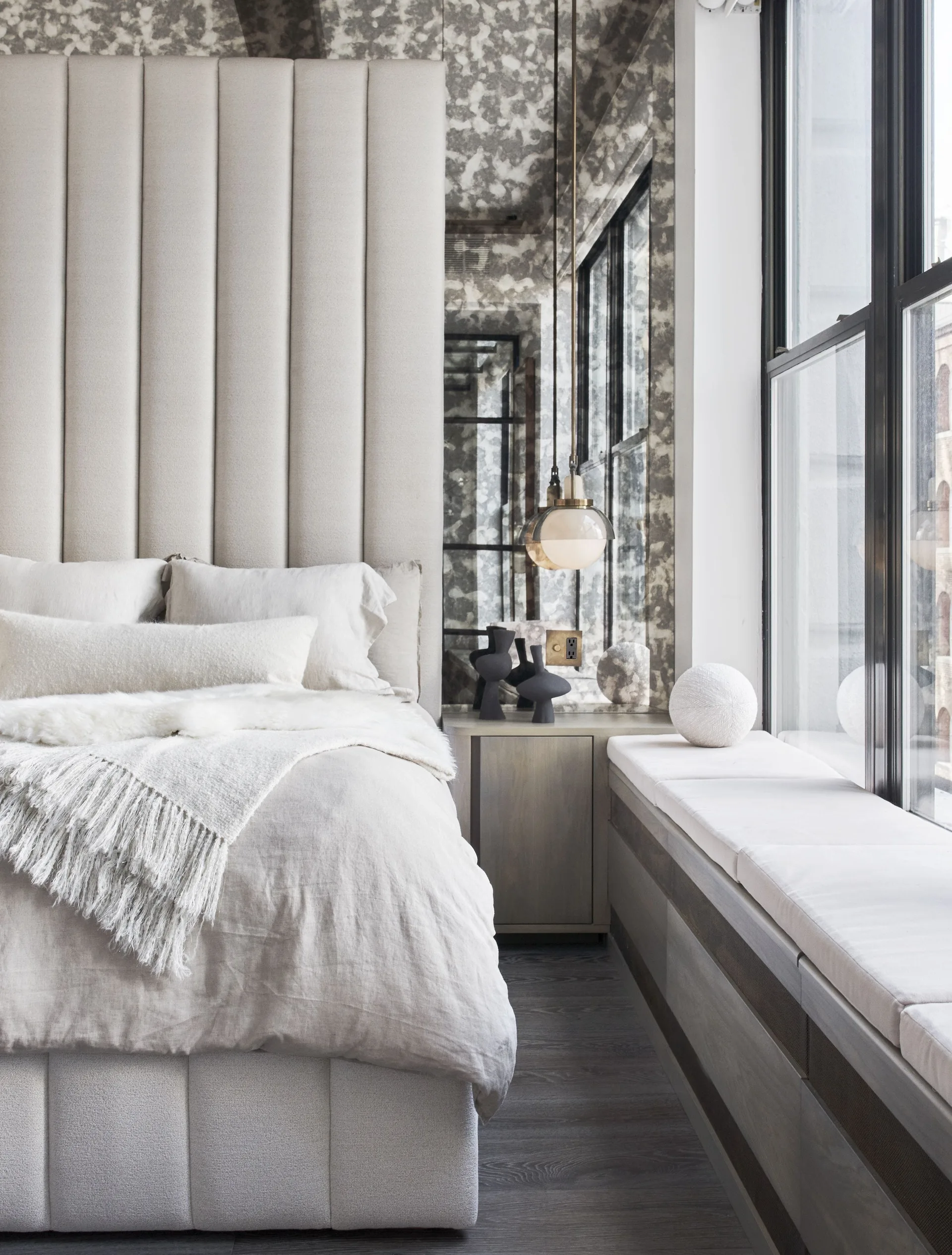 A contemporary bedroom featuring a tall channel-tufted headboard, neutral bedding, a built-in window bench, and antiqued mirrored walls.