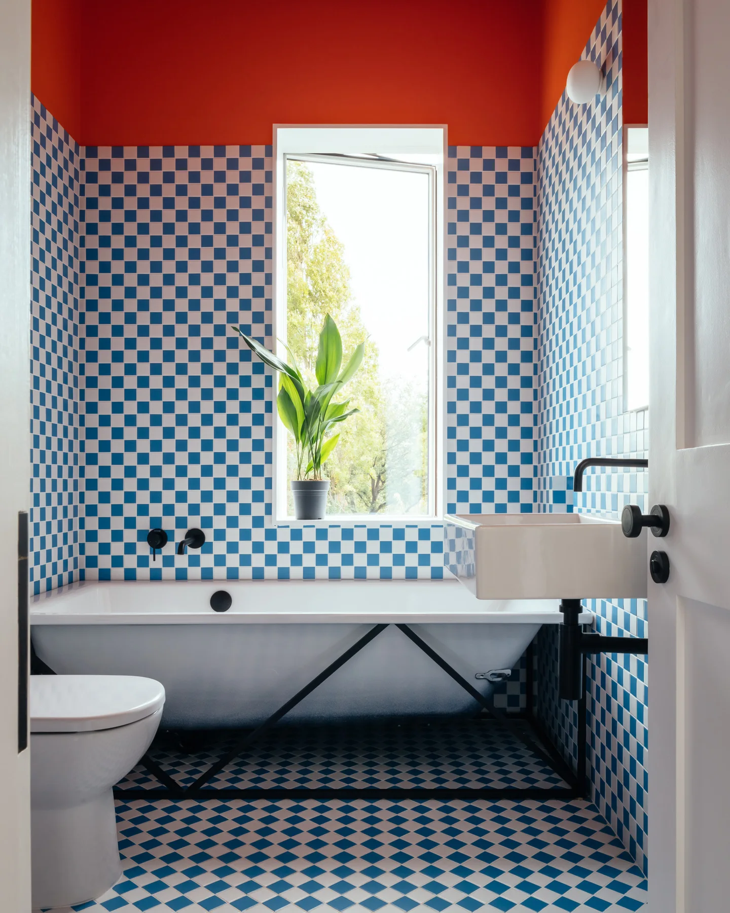 A modern bathroom featuring blue and white checkered tile walls and floors, a white bathtub under a window, and a bright orange ceiling.