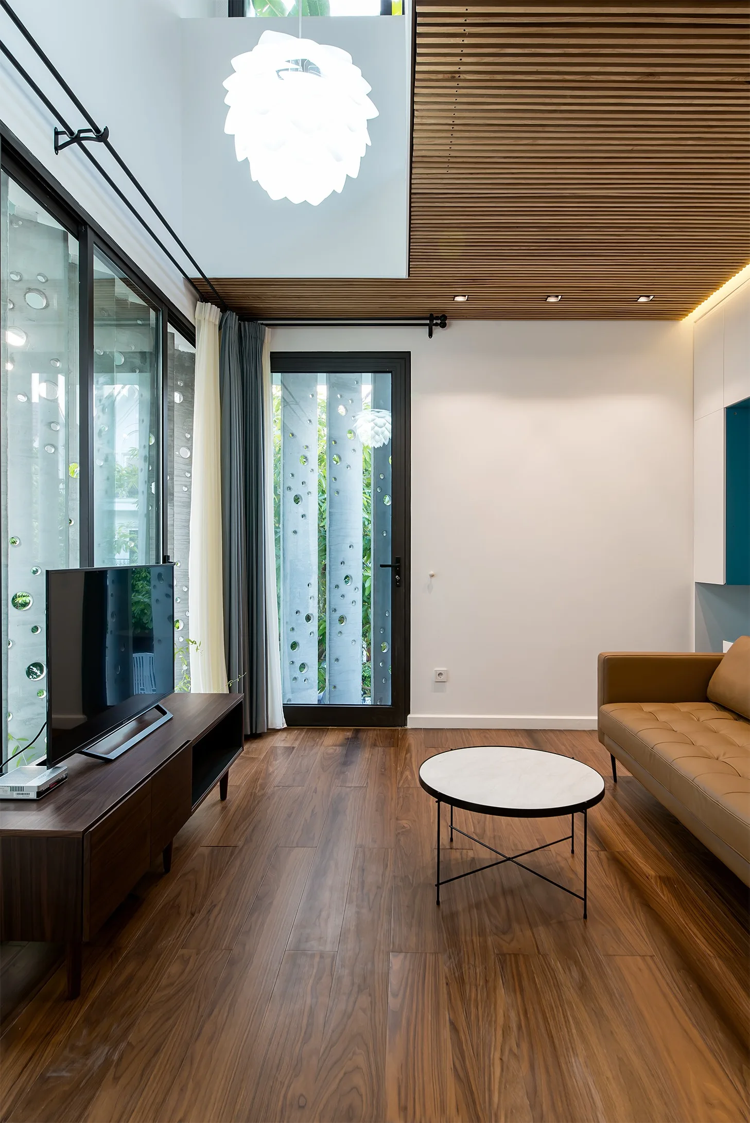 A modern living room features a tan leather sofa, a round marble coffee table, dark wood floors, and a distinctive linear wood slat ceiling.