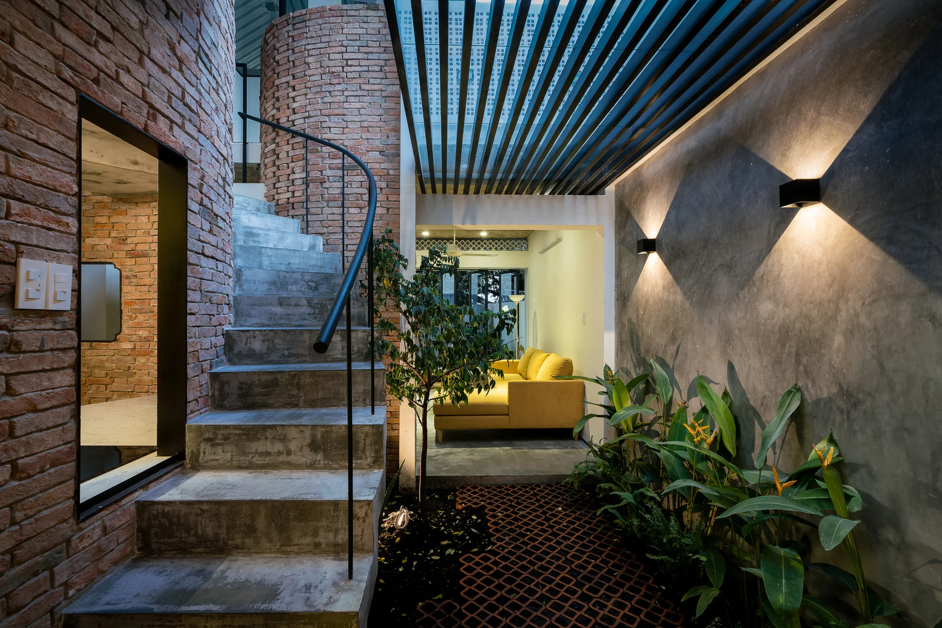 An indoor courtyard features a curved concrete staircase beside a brick wall, opposite a gray plaster wall decorated with plants and wall sconces.