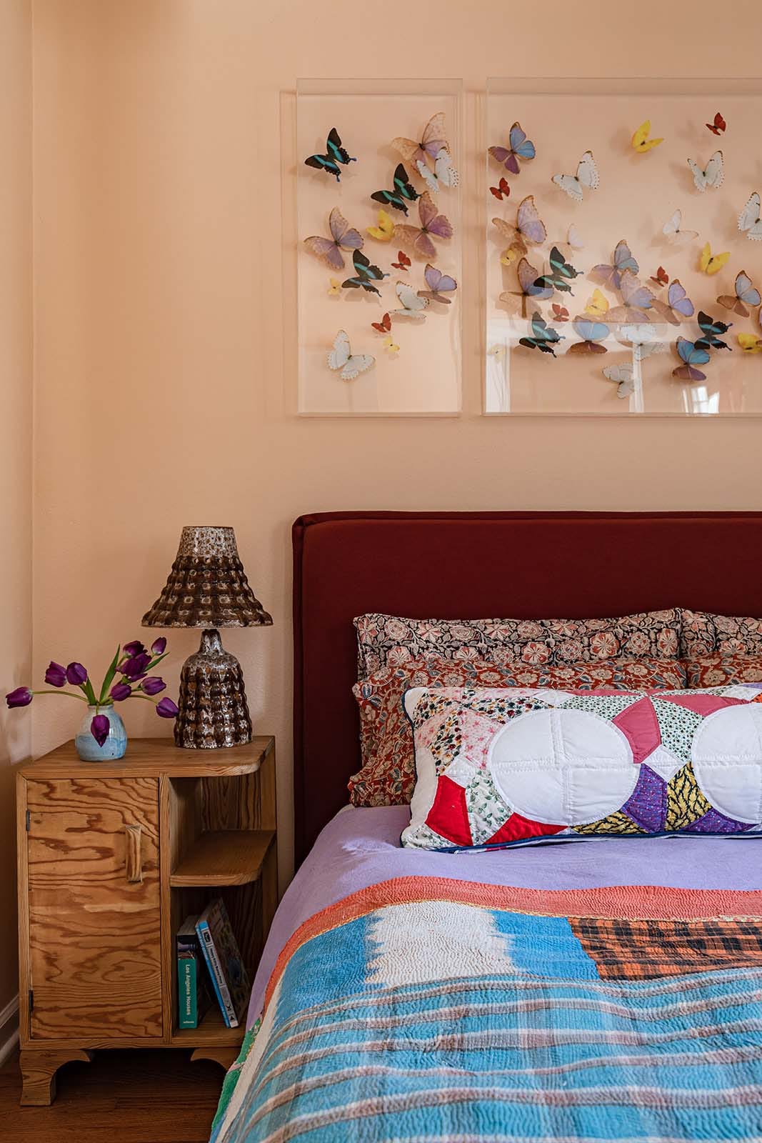 A bedroom corner features a burgundy headboard against peach walls, decorated with patterned bedding, a wooden nightstand, and framed butterfly art.