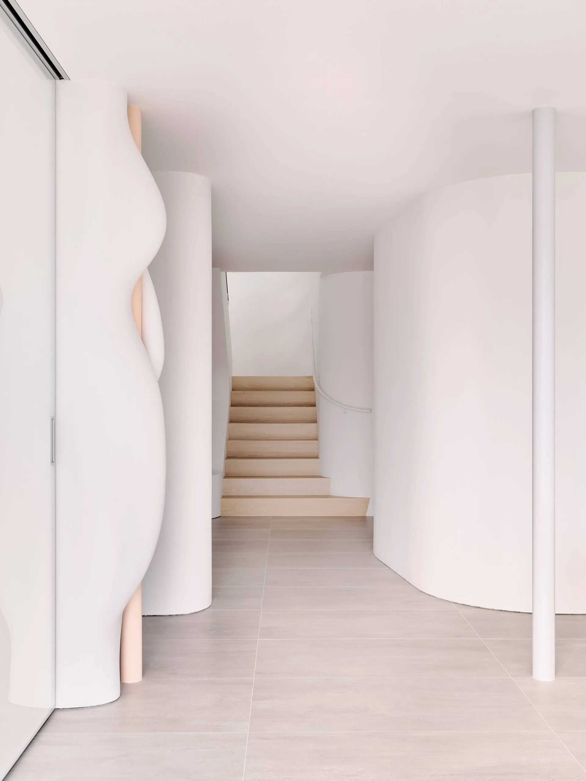 A minimalist hallway featuring curved white walls, large floor tiles, and a light wood staircase in the background.