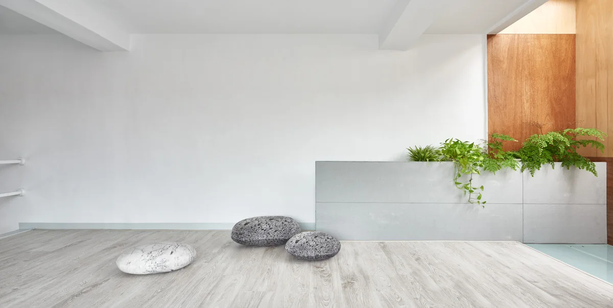 A minimalist white room features light wood floors, a large grey built-in planter with green plants, and three stone-shaped floor cushions.