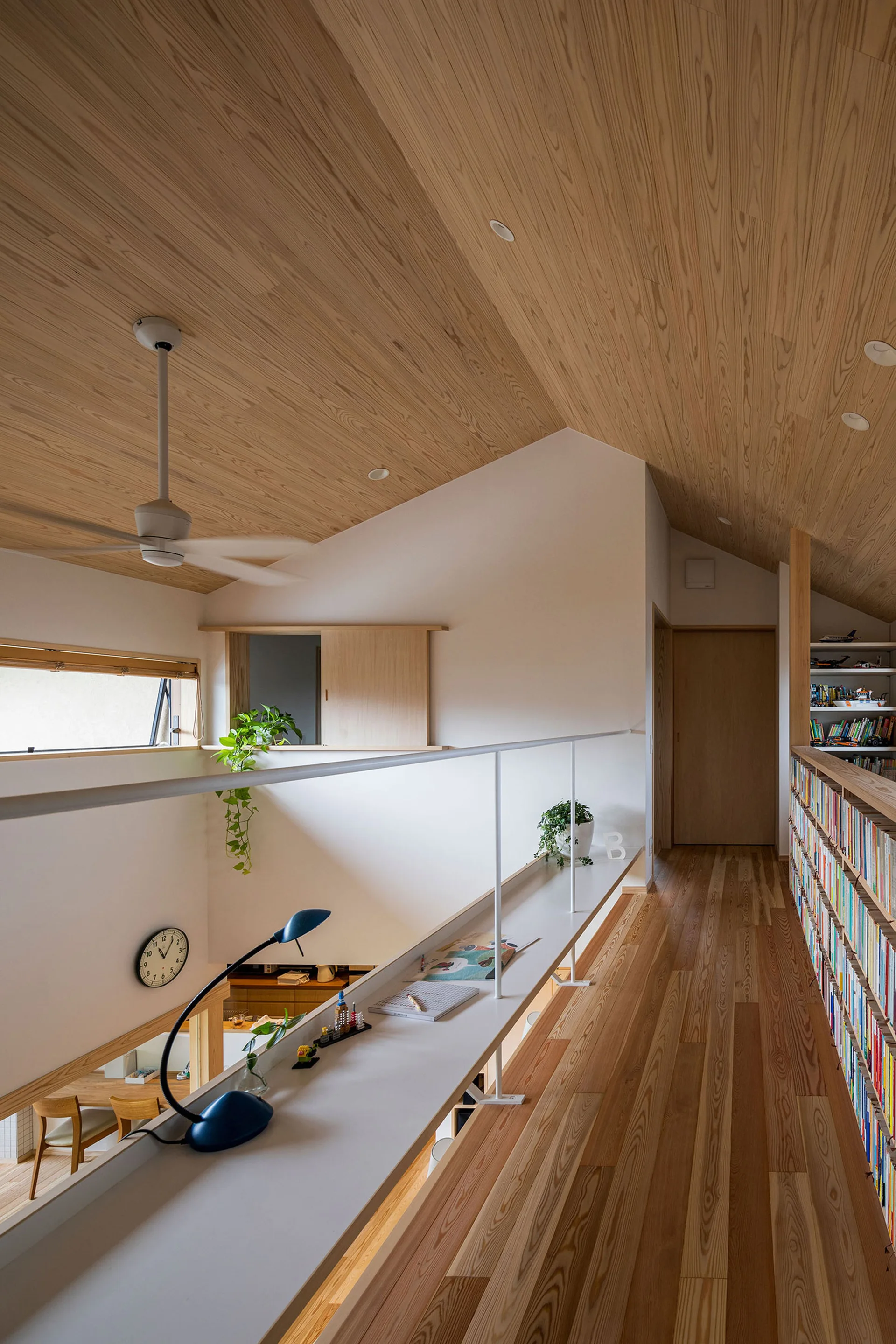 A mezzanine landing features a sloped wood plank ceiling, a long white desk integrated with the railing, and a low bookcase filled with books.
