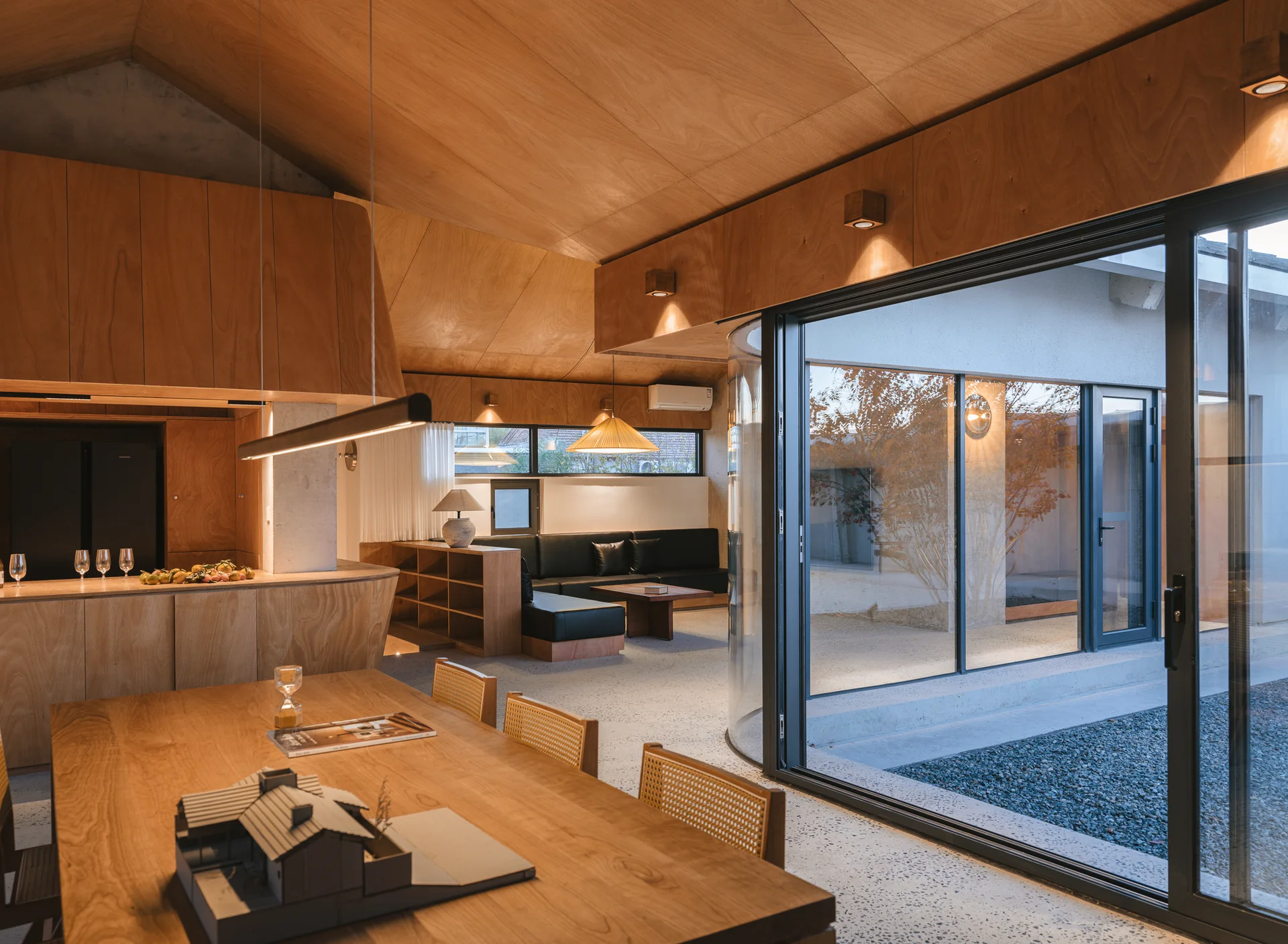A modern open-plan dining and living space features extensive warm wood paneling, a large curved glass wall, terrazzo floors, and a black leather sofa.