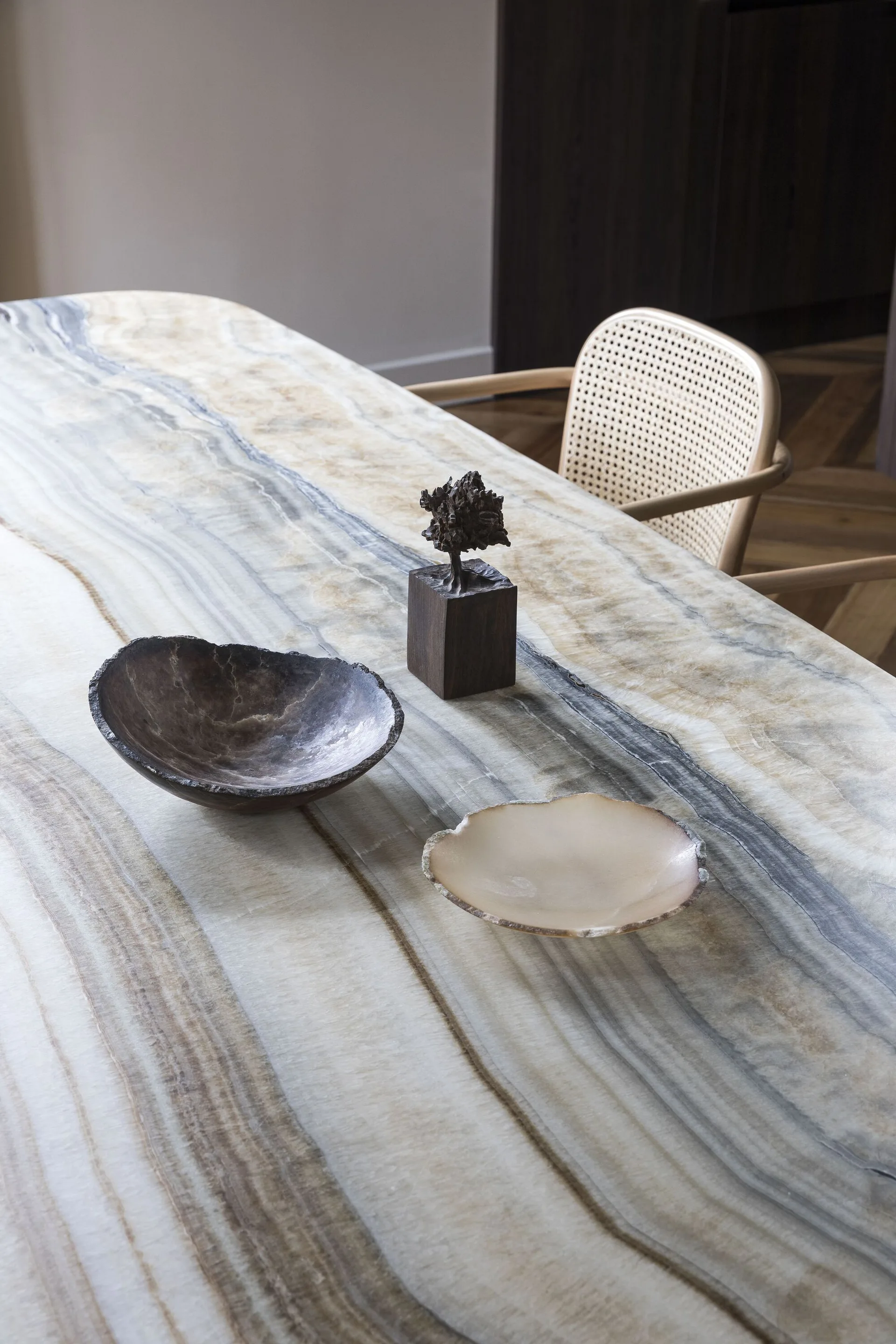 A natural veined stone dining table topped with organic decorative bowls and a small sculpture, set beside a woven cane chair.