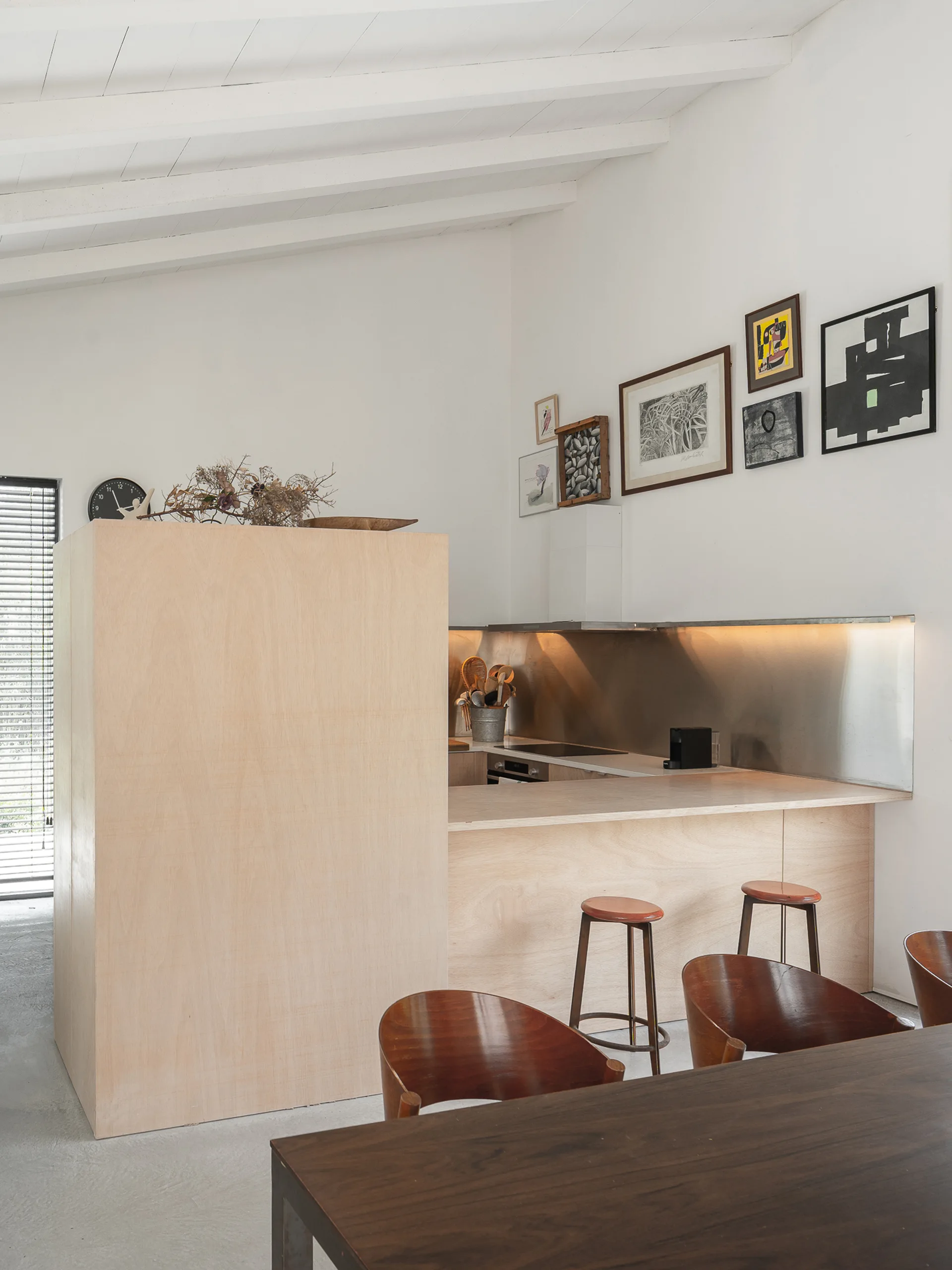 A minimalist kitchen features a light plywood island, bar stools, a metal backsplash, and a small gallery wall under a vaulted ceiling.