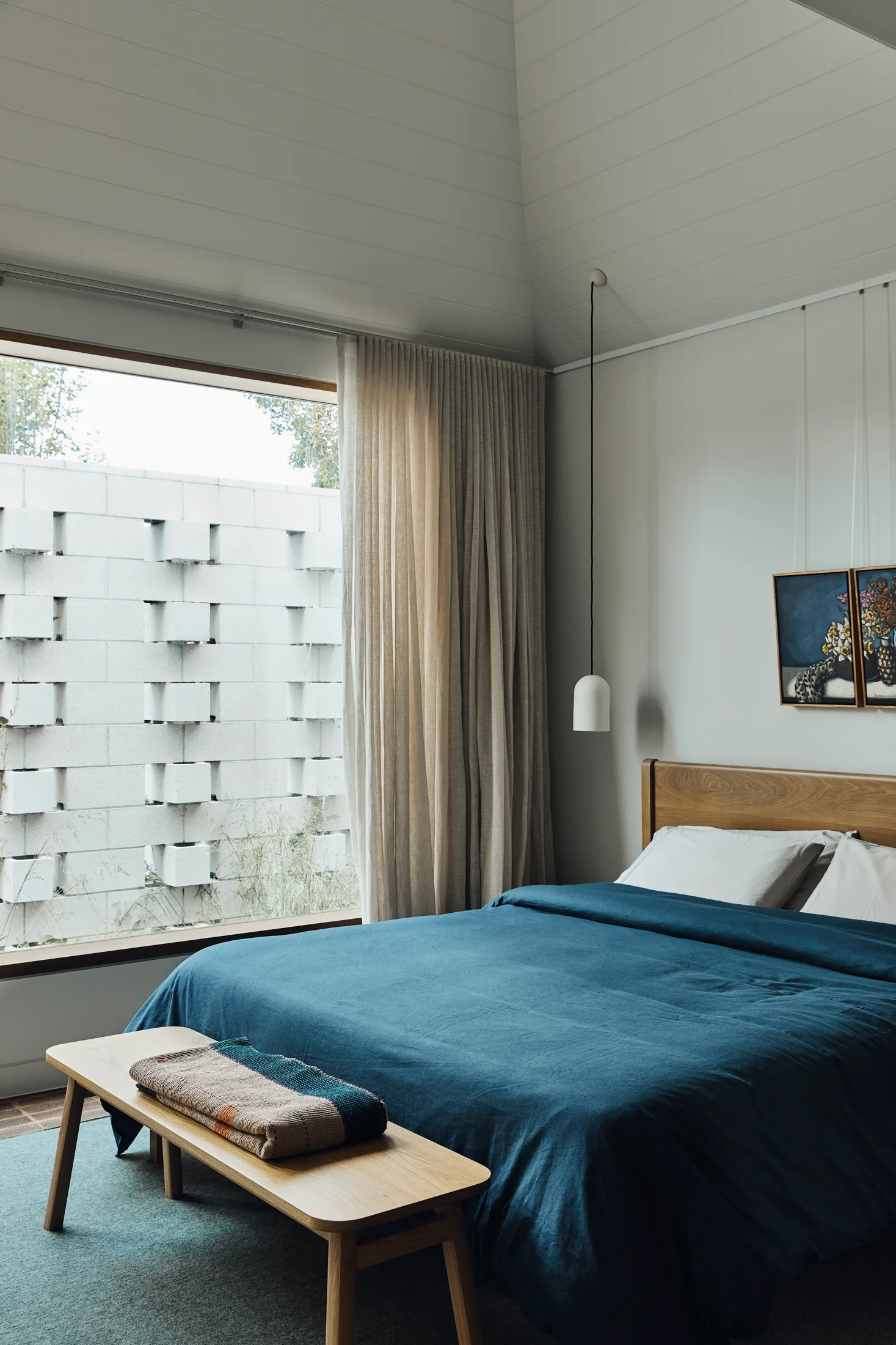 A modern bedroom featuring a wood bed with a blue duvet, a matching wooden bench, and a large window framing a patterned concrete block wall.