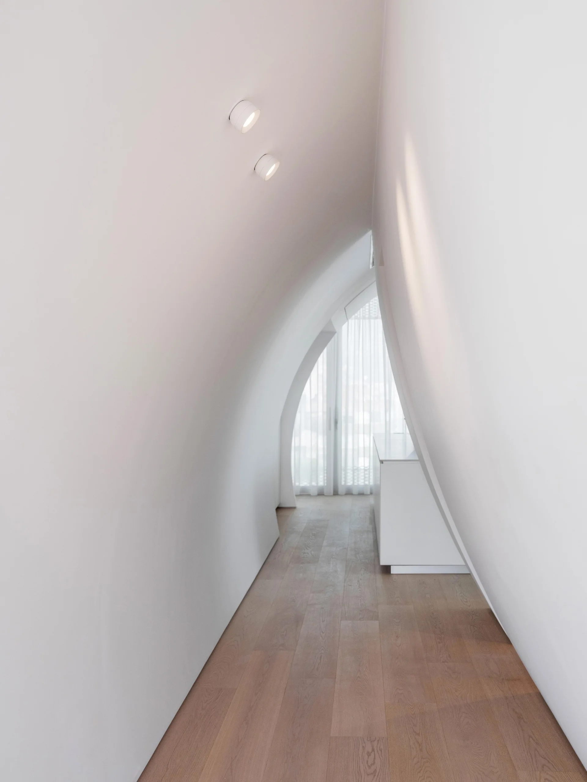 A minimalist hallway featuring curved white plaster walls, light wood flooring, and a window with sheer white curtains.