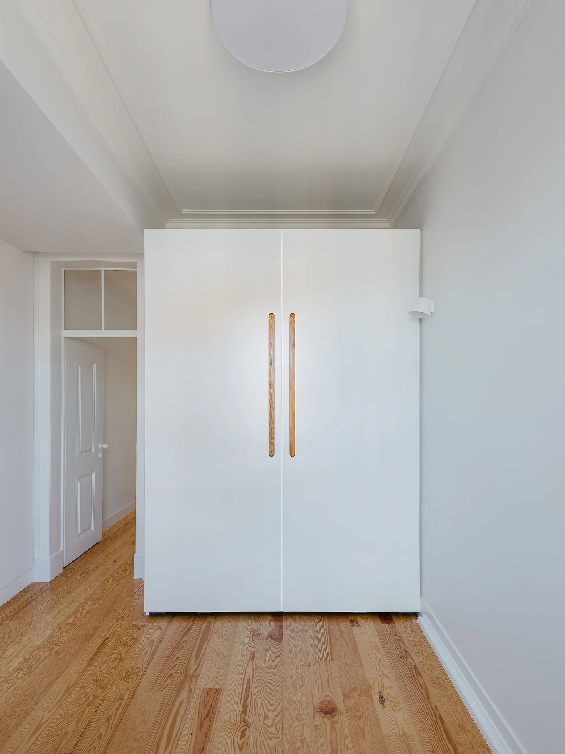 A minimalist white floor-to-ceiling closet with long vertical wooden handles stands on a light pine wood floor in a bright hallway.