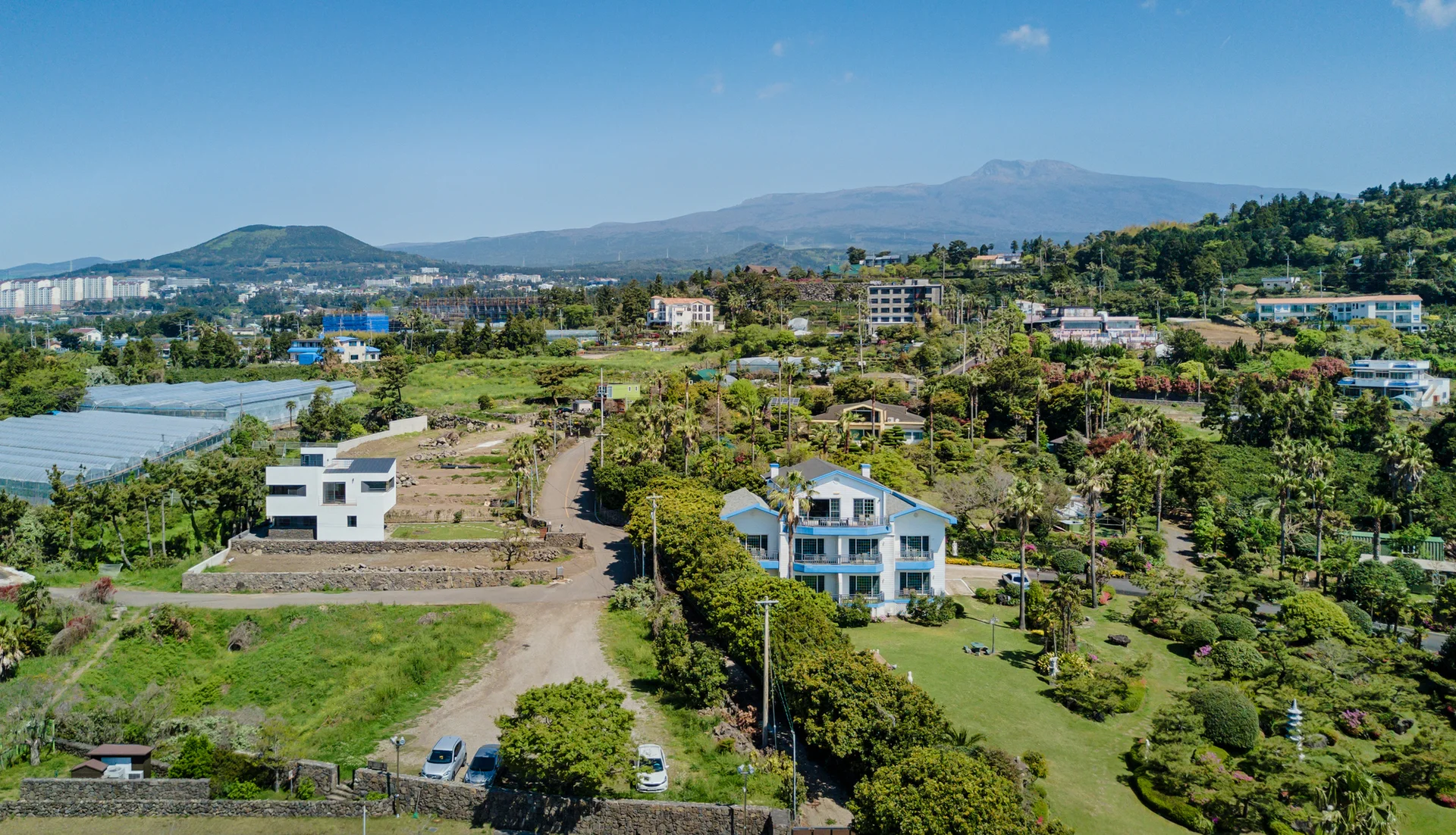 An aerial view of a hillside landscape featuring white stucco homes, dark volcanic stone walls, palm trees, and a mountain in the distance.
