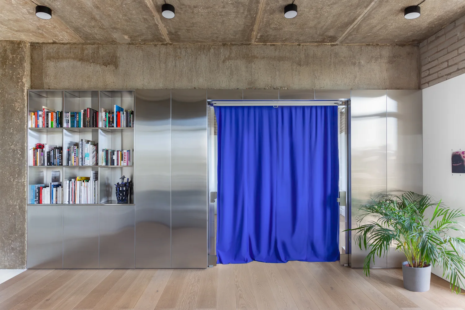 A modern living room features a full-height brushed metal wall unit with built-in bookshelves and a bright blue fabric curtain divider.