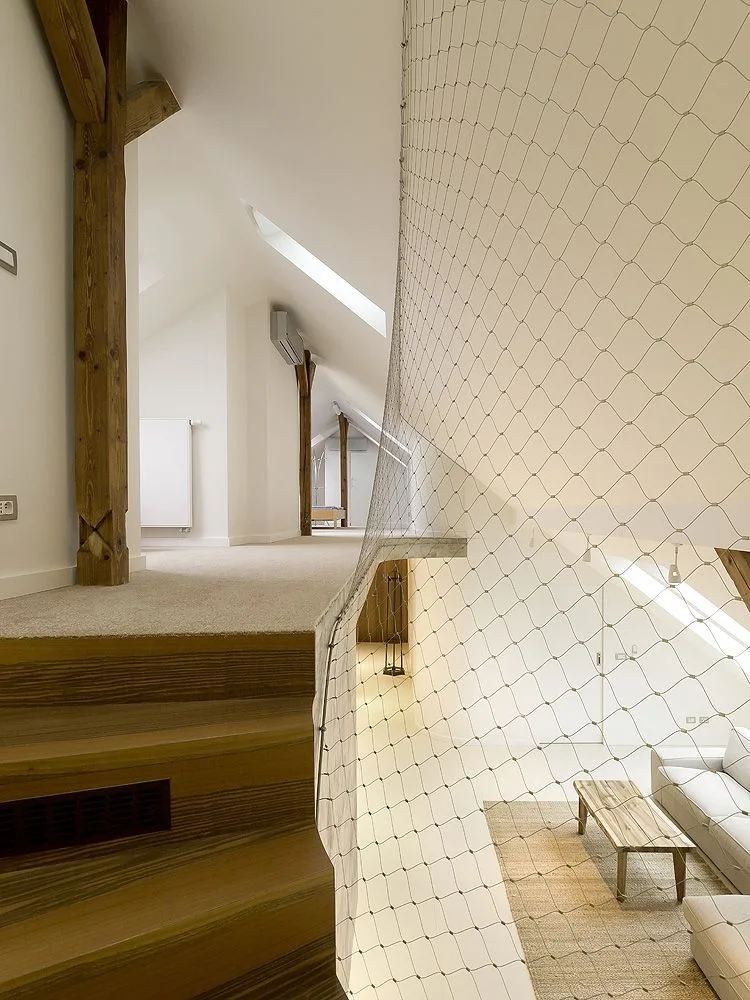A modern loft mezzanine with a white safety net barrier overlooks a living room below, featuring exposed wooden beams and a carpeted landing.