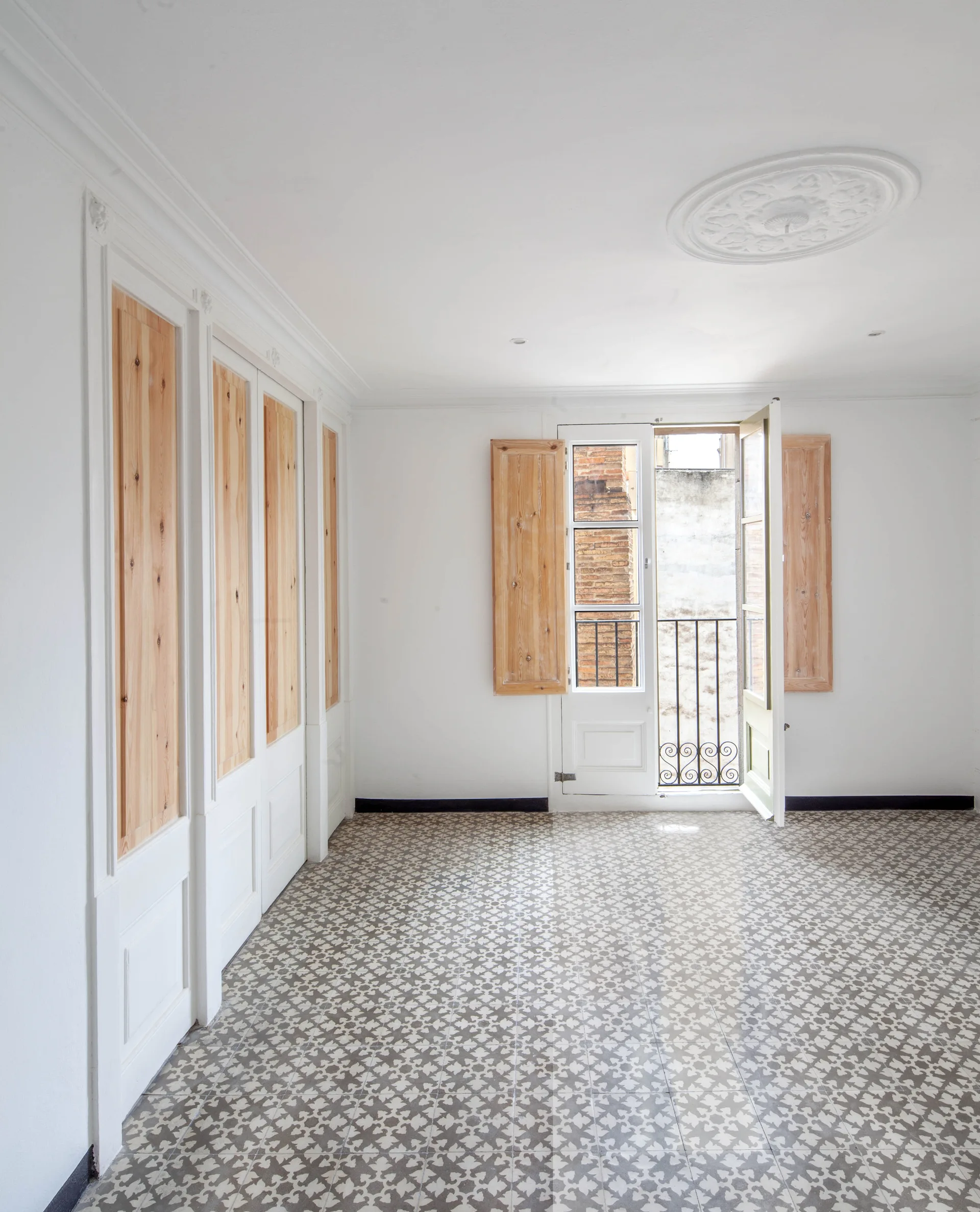 An empty room features gray and white patterned floor tiles, tall white built-in closets with natural wood panels, and matching wood shutters.