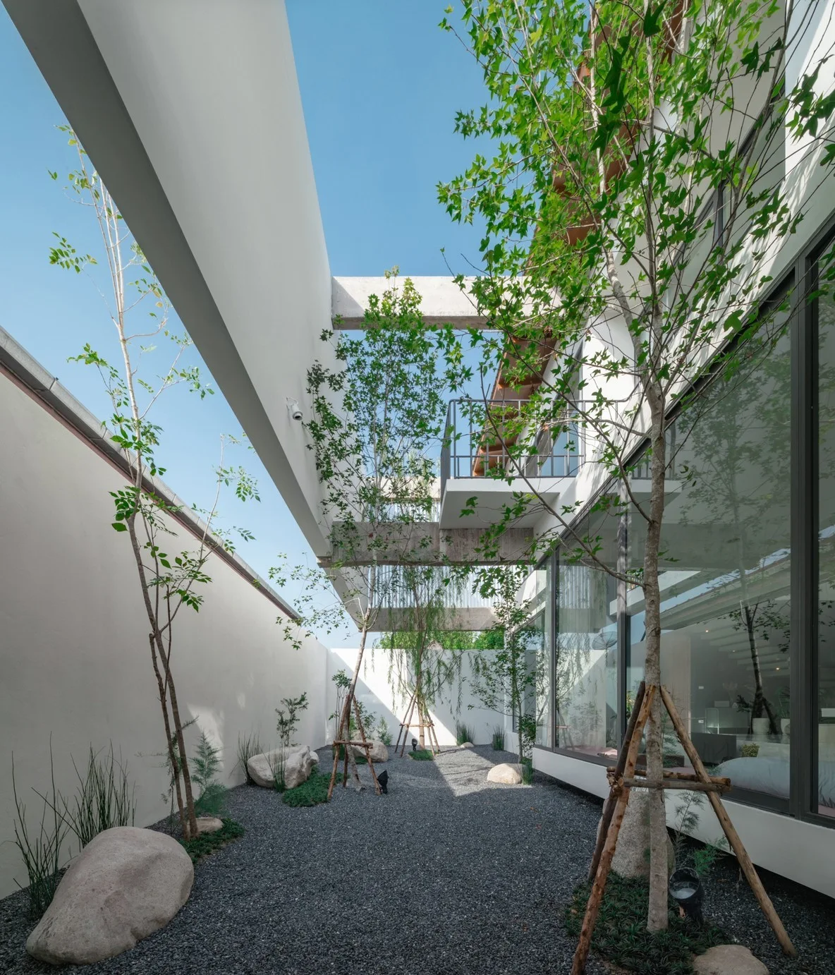 A modern courtyard features a dark grey gravel ground, young trees, white walls, and overhead concrete beams under a clear blue sky.
