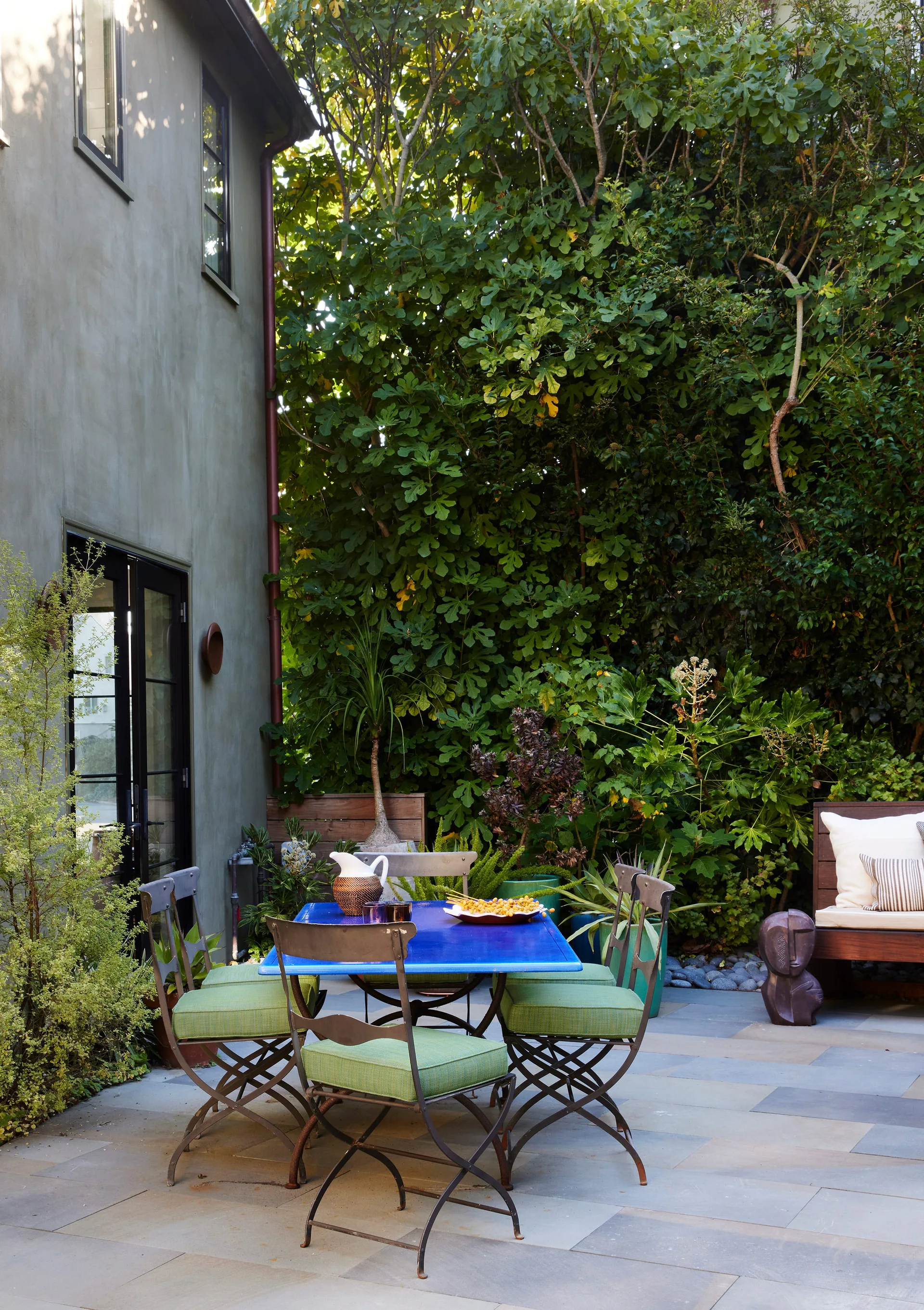 A stone tile patio features a bright blue dining table with green-cushioned metal chairs, set against a backdrop of dense green trees and a stucco wall.