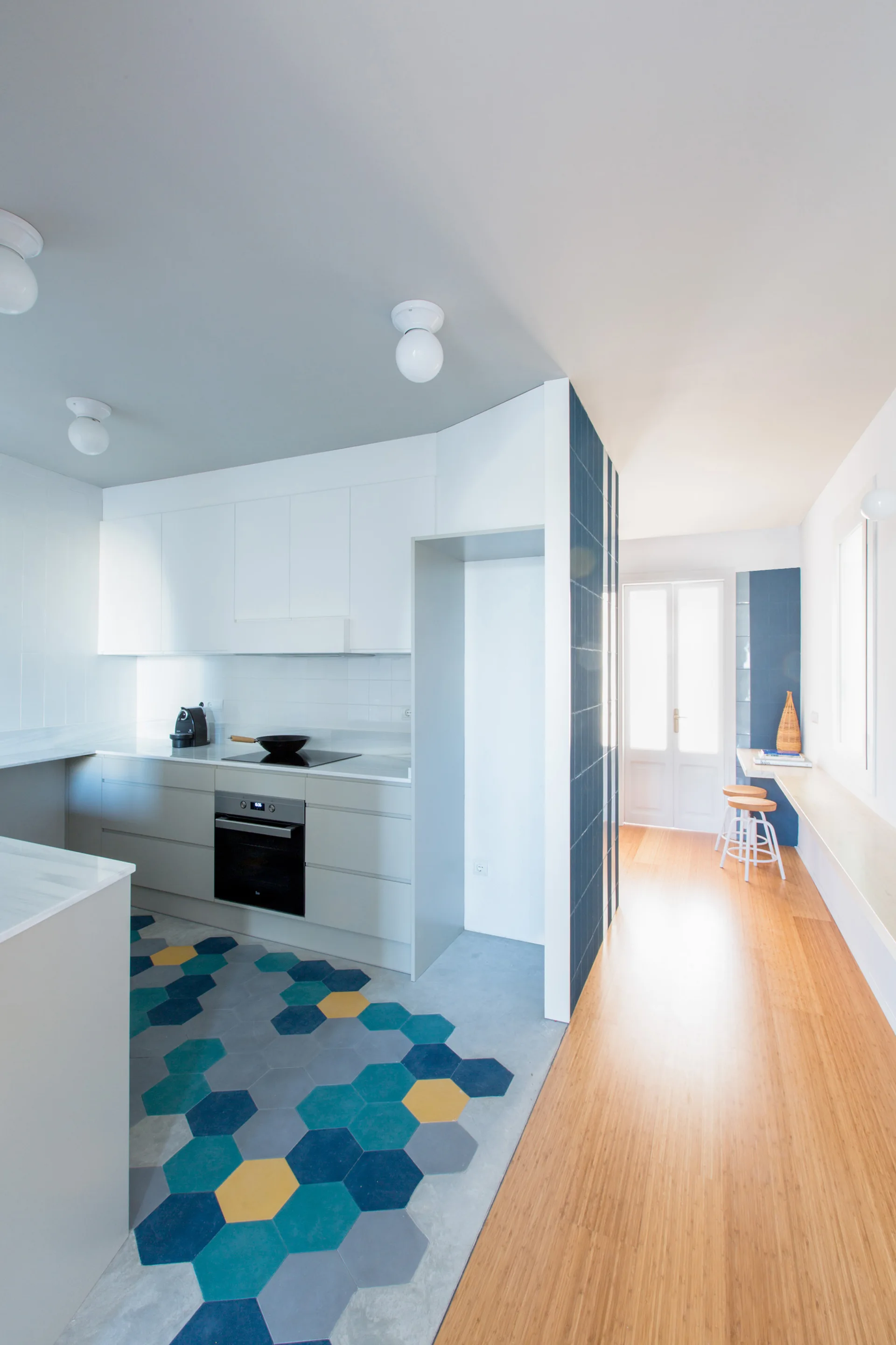 A kitchen with grey cabinets and blue hexagonal floor tiles transitions into a hallway with a long wooden desk and wood flooring.
