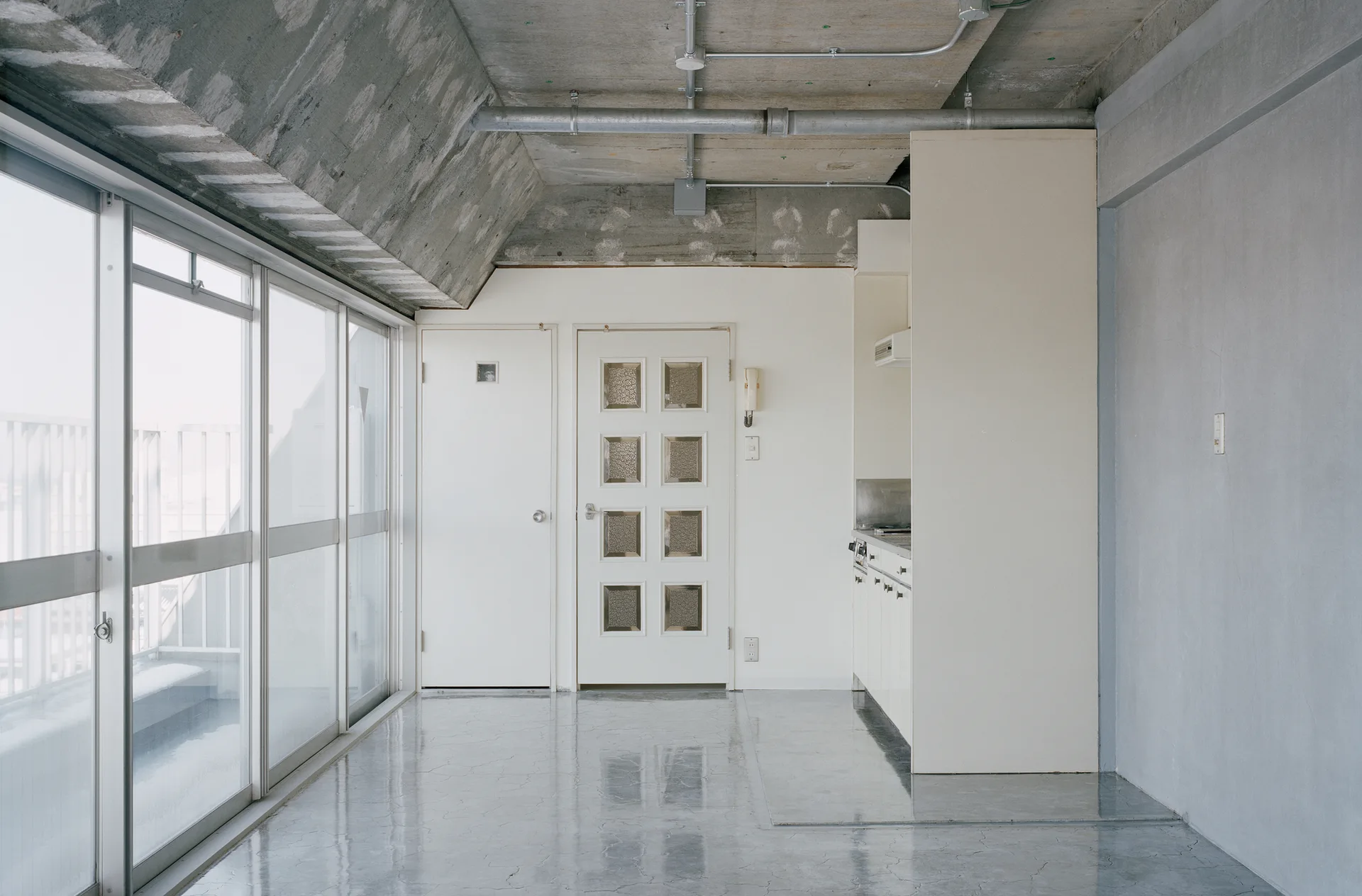 A minimalist industrial room featuring a polished concrete floor, raw concrete ceiling with exposed pipes, white doors, and large sliding glass windows.