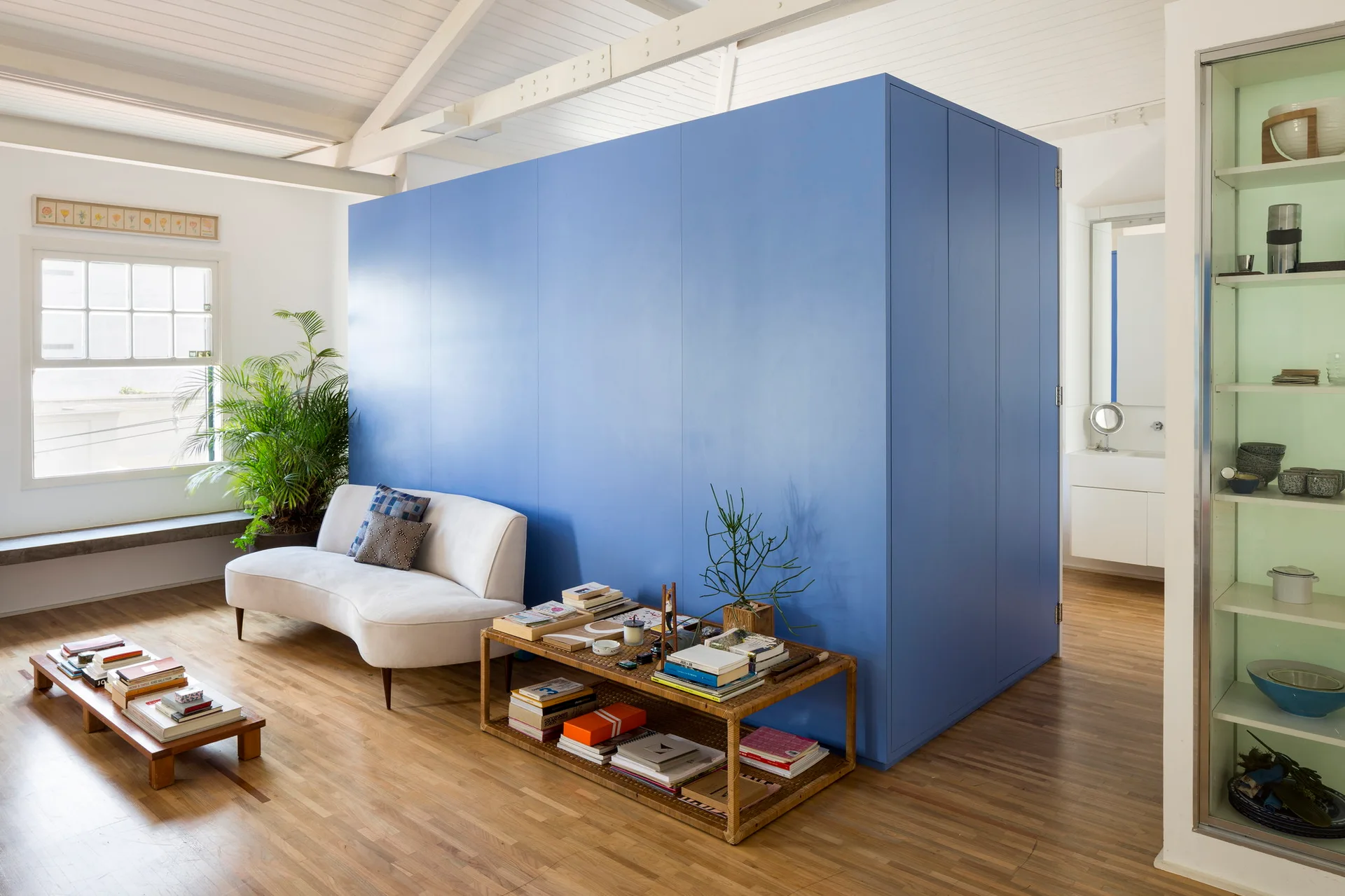 A bright living room features a large blue central partition wall, a curved white sofa, a low wooden coffee table, and exposed white ceiling rafters.