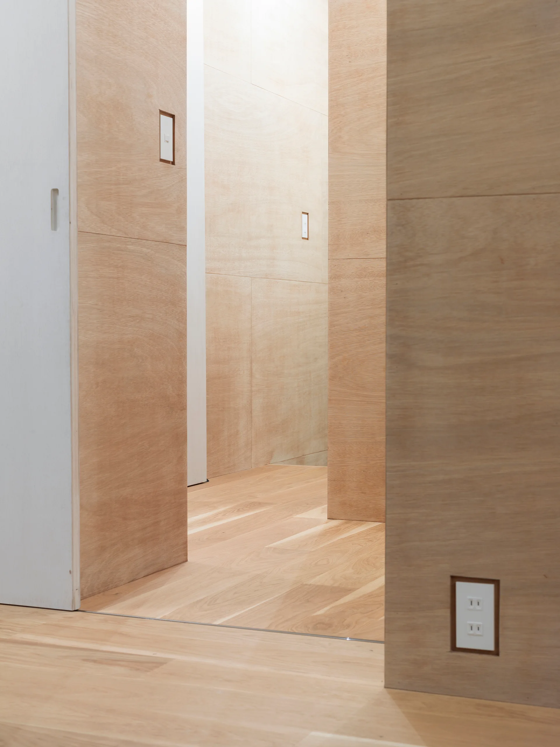 A minimalist hallway features light wood paneled walls, matching plank flooring, a white sliding door, and recessed electrical outlets and switches.