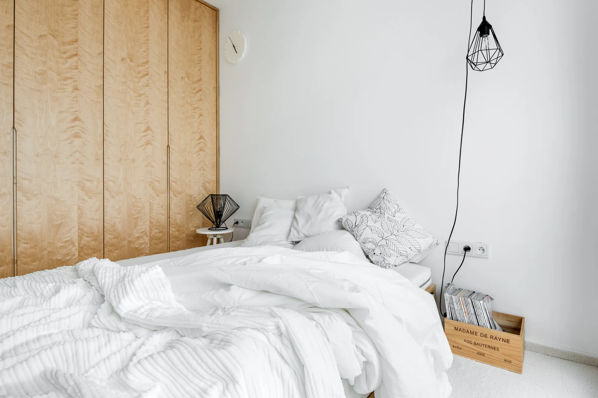 A minimalist bedroom displays a large light wood wardrobe alongside a bed with white textured bedding and black geometric lighting.