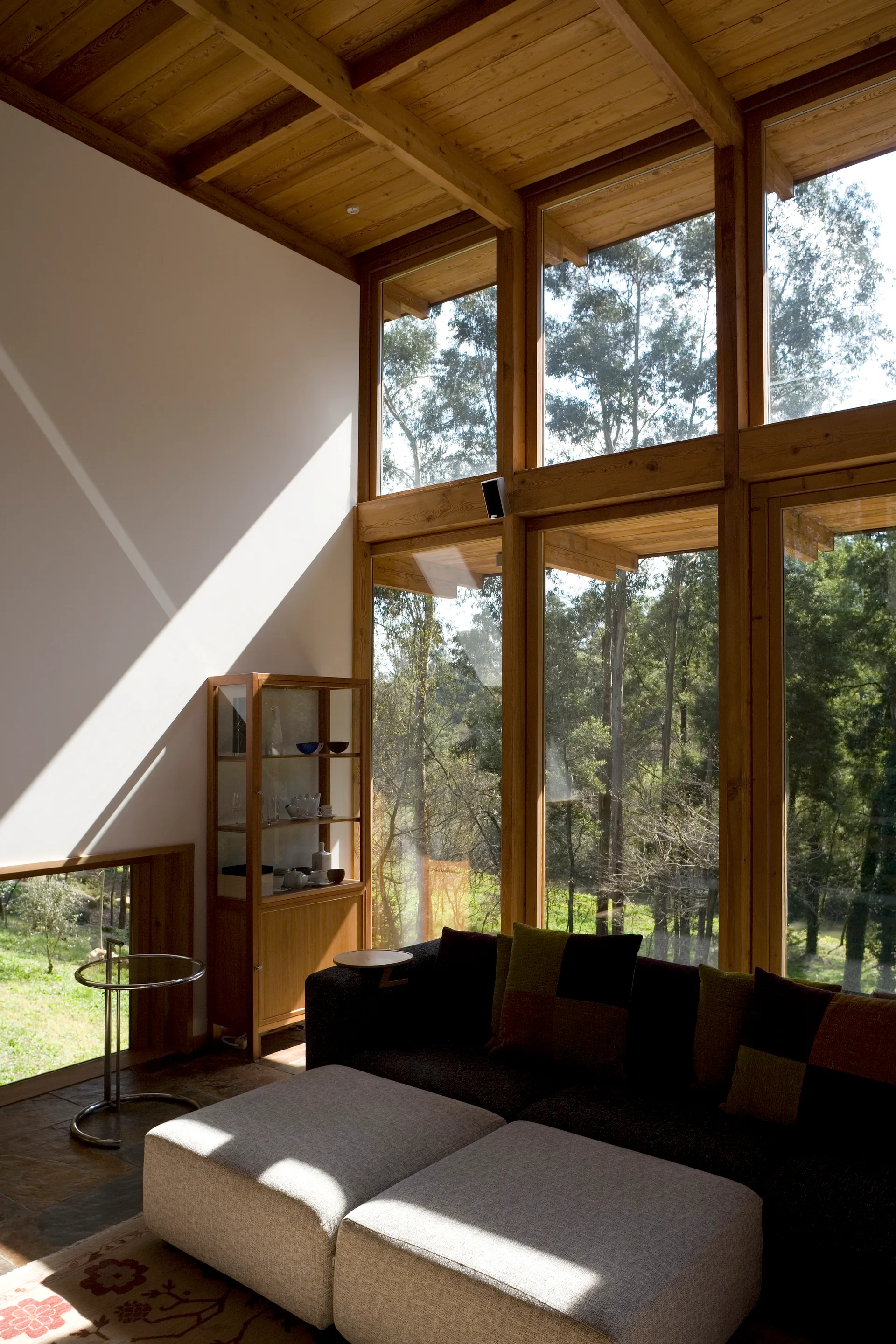 A modern living room features a dark sofa, light-colored ottomans, and a wood display cabinet next to large floor-to-ceiling windows showing forest views.