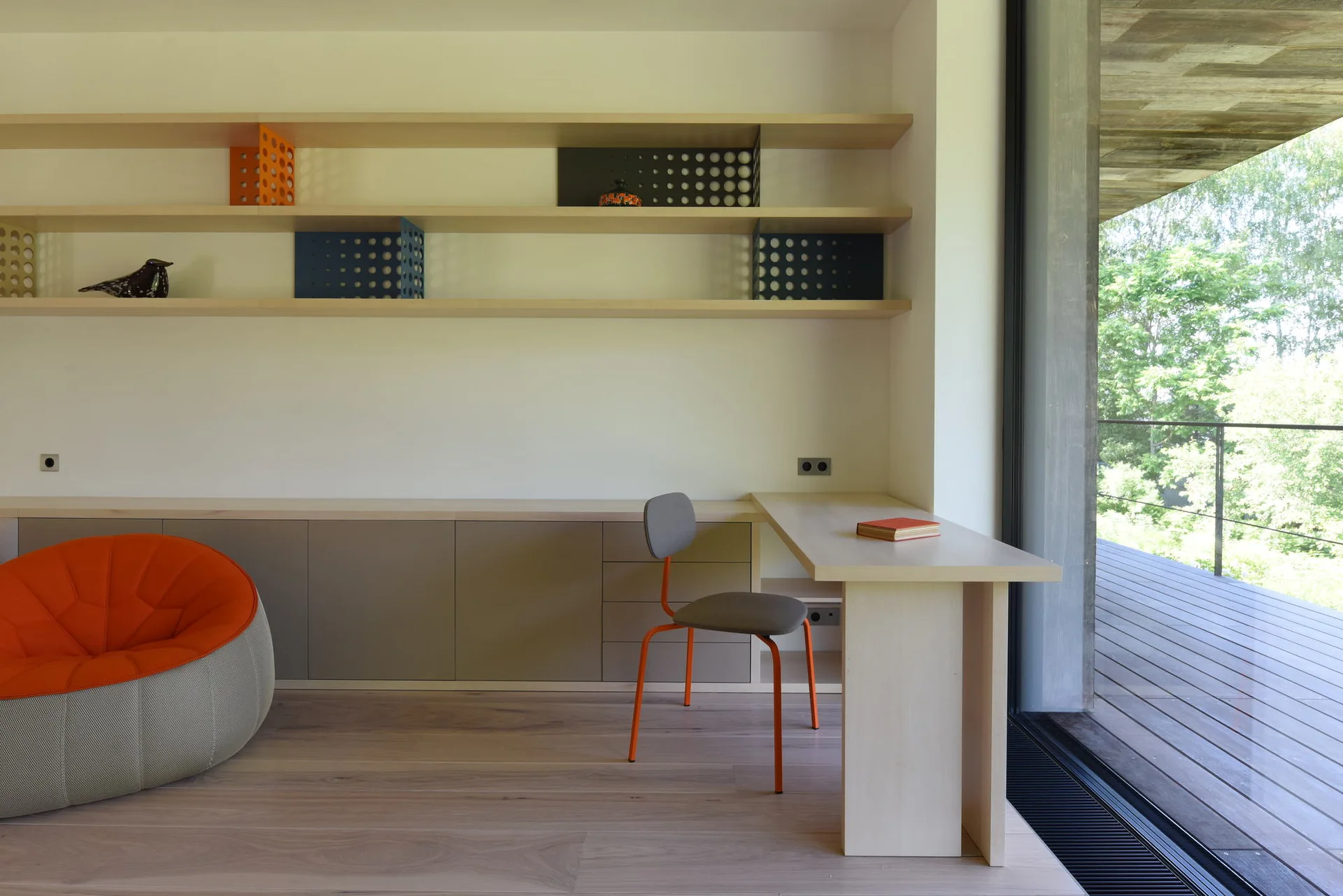 A minimalist home office features a built-in light wood desk, floating shelves, a grey chair with orange legs, and a large window opening to a deck.