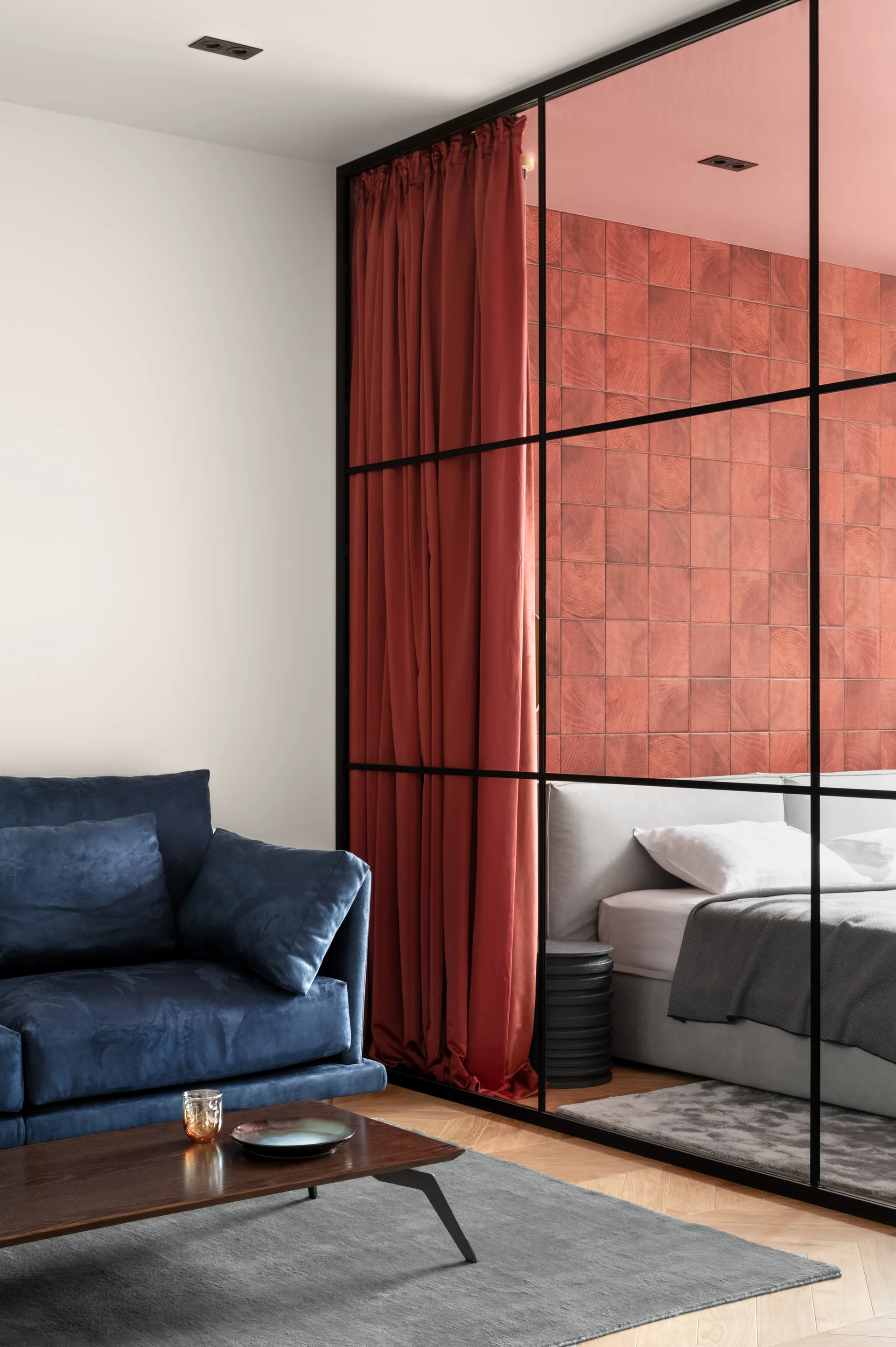 A blue sofa in a living room sits next to a black metal-framed glass partition looking into a bedroom with a red tiled wall and matching curtain.