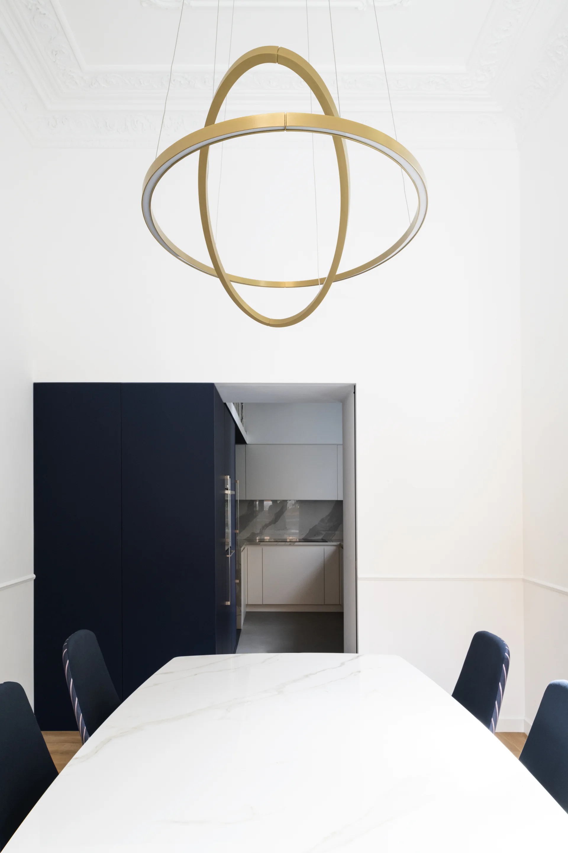 A modern dining room features a white marble table, navy blue chairs, and a large gold circular pendant light hanging from a decorative molded ceiling.