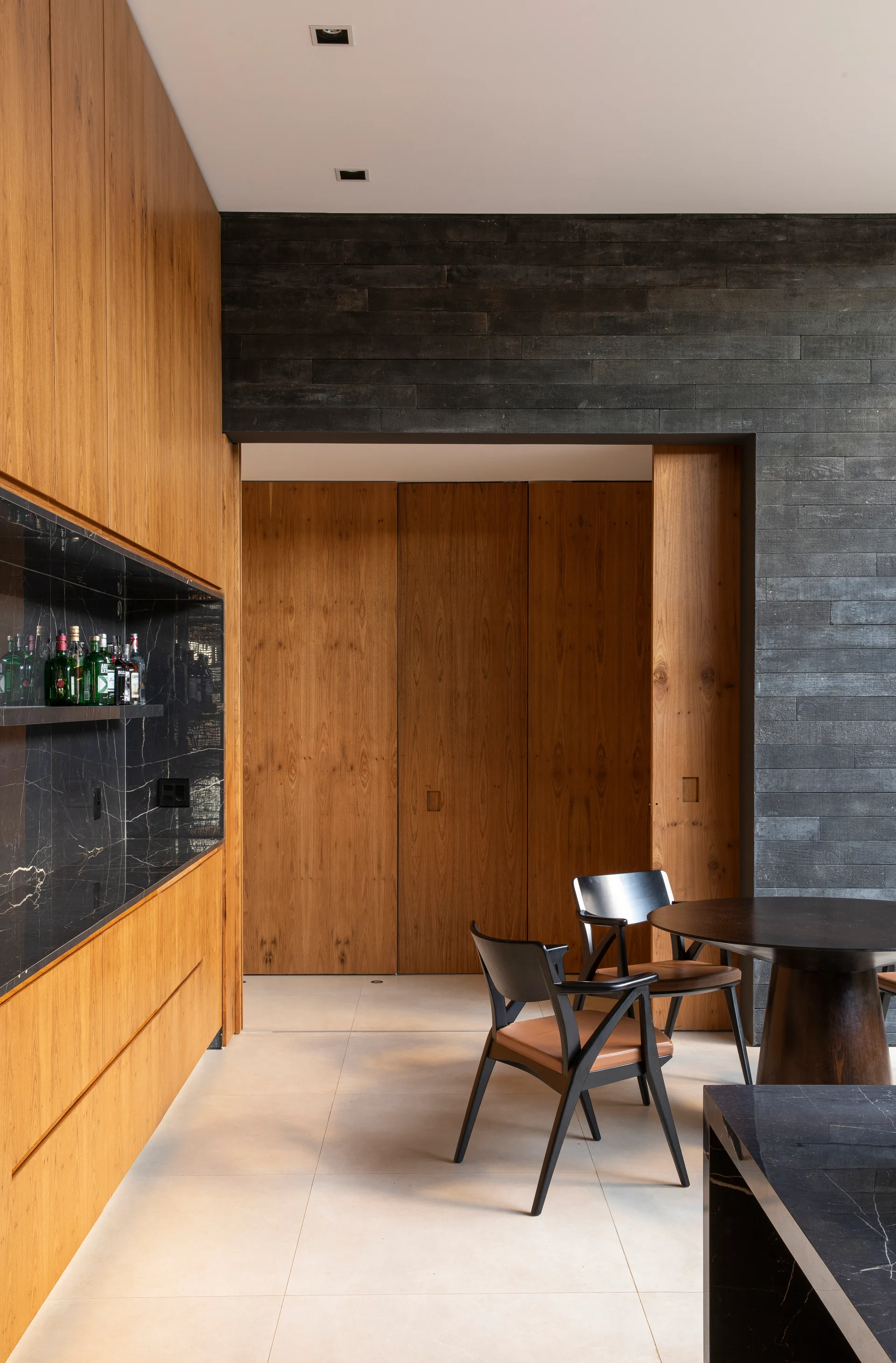 A modern home bar features warm wood cabinetry, a black marble backsplash, and a dining area with a round dark wood table and black chairs.