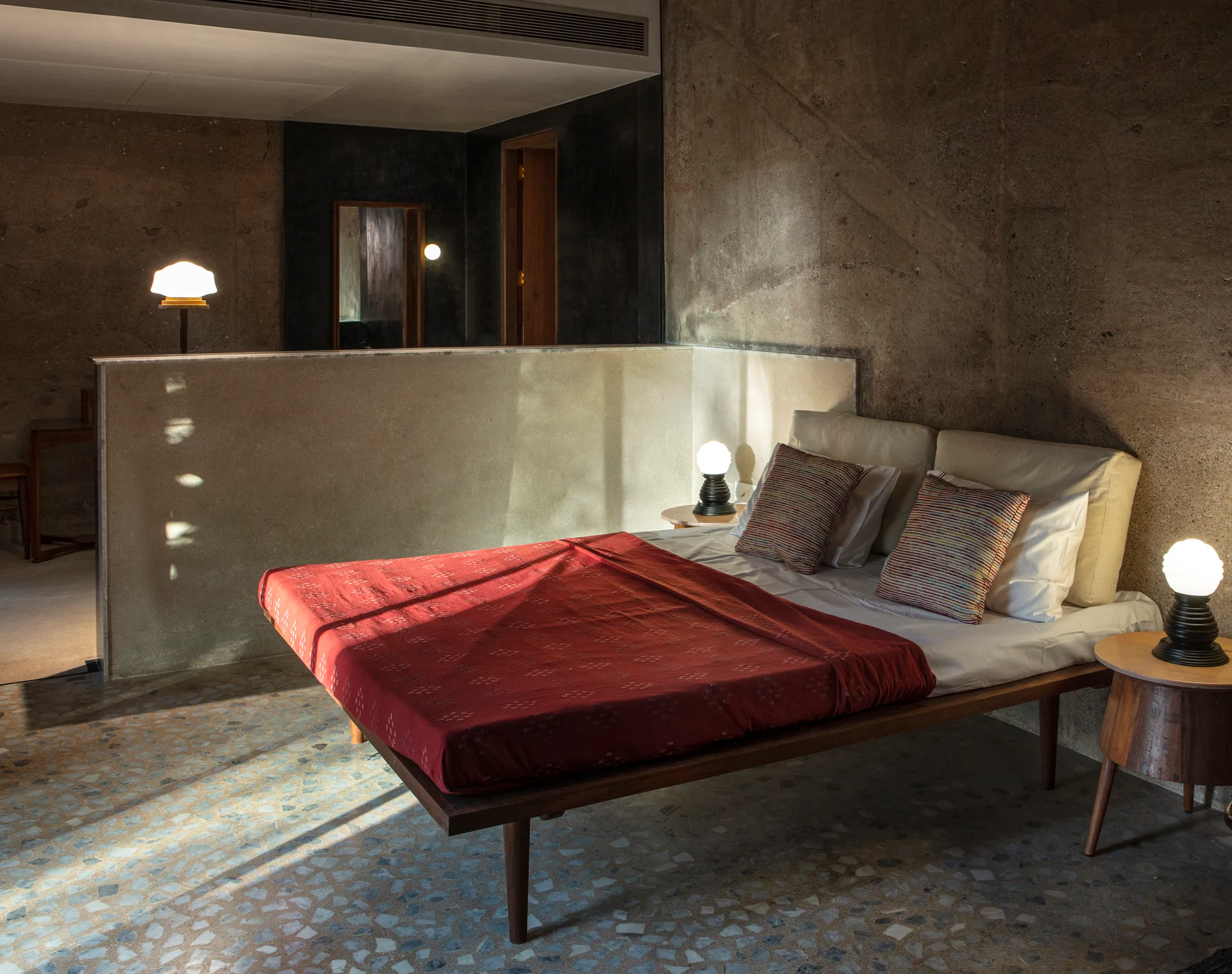 A low-profile wooden bed with a red coverlet sits in a concrete bedroom with speckled terrazzo floors, flanked by round bedside tables.