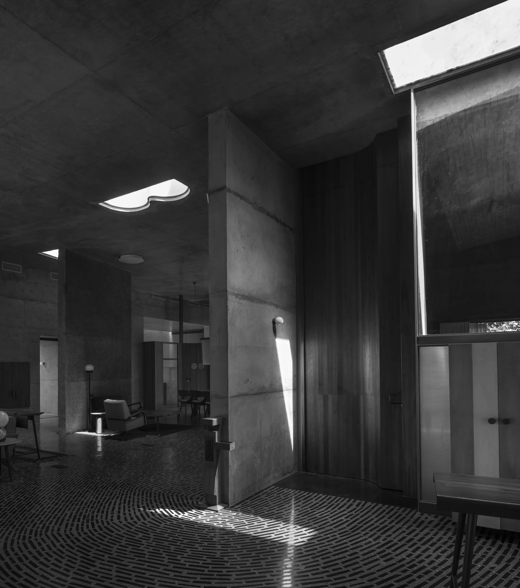 A black and white photo of a brutalist interior with raw concrete walls, a radial patterned brick floor, and architectural skylights.