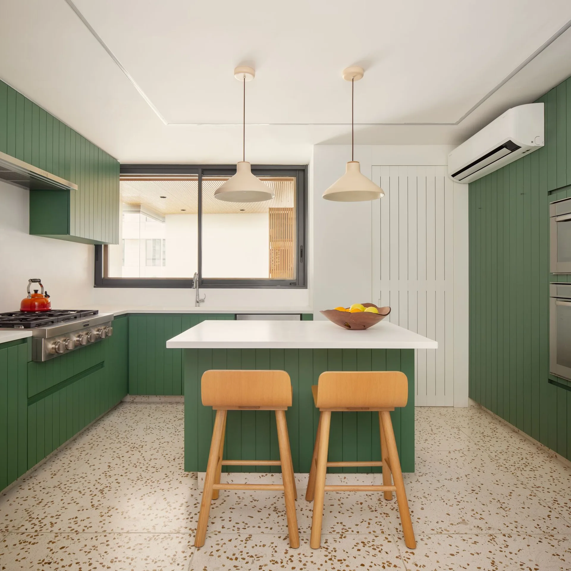 A modern kitchen featuring green vertical-grooved cabinets, a white-topped kitchen island with wood bar stools, and speckled terrazzo flooring.