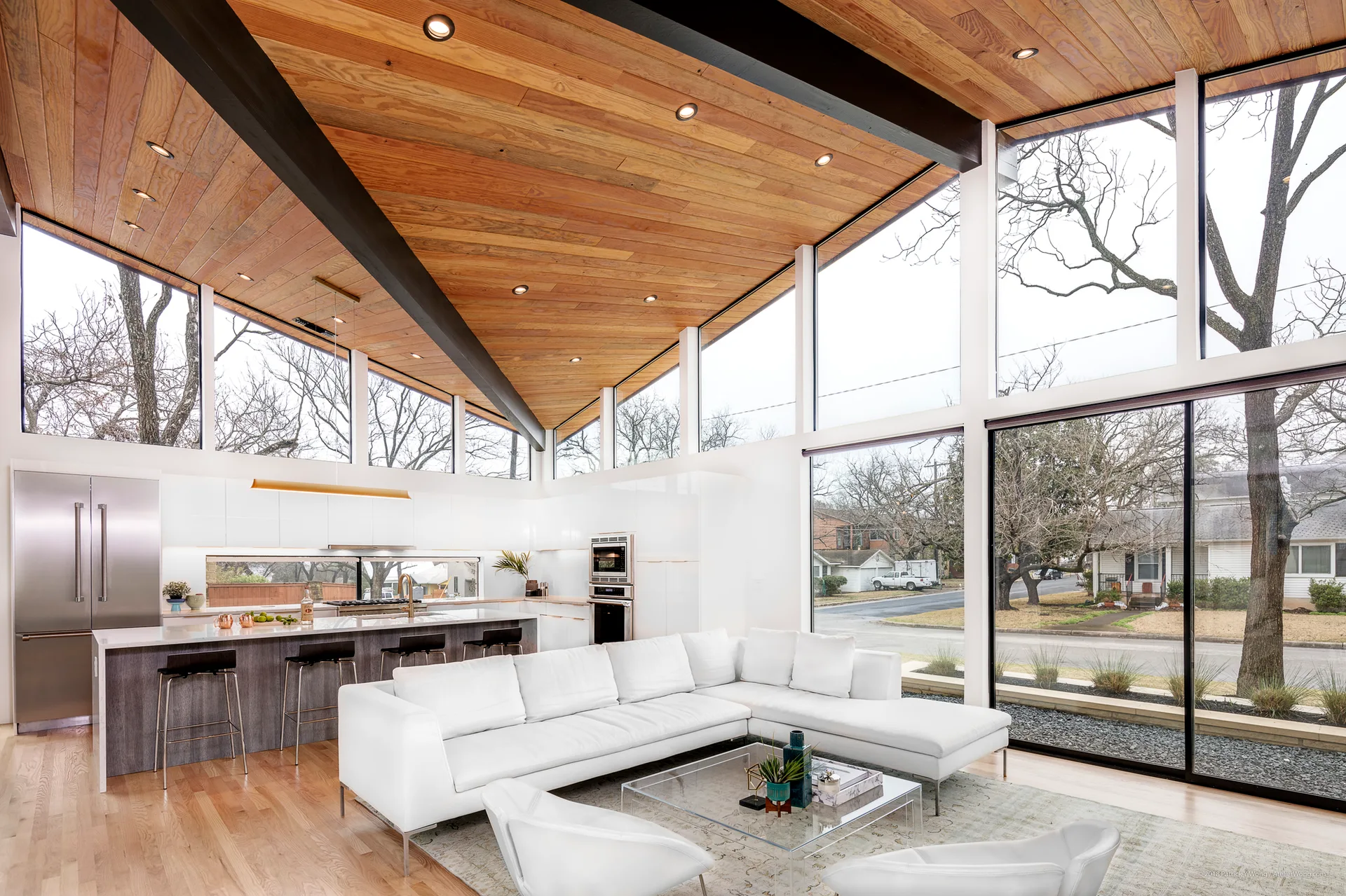 A bright open-plan living room and kitchen featuring a vaulted wood plank ceiling, a white sectional sofa, and expansive glass window walls.