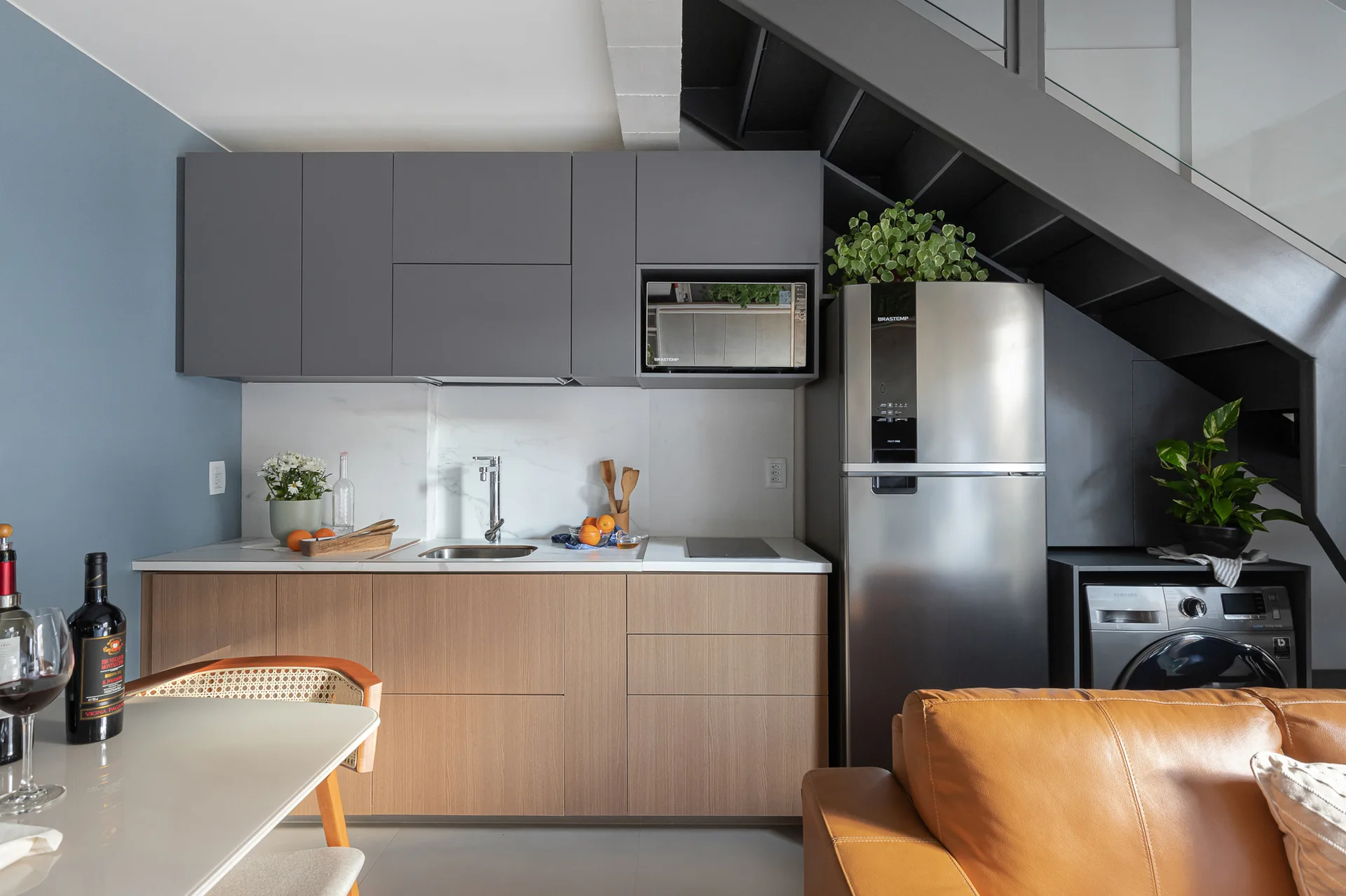 A modern kitchen featuring grey and wood cabinets, a stainless steel refrigerator, and a washing machine tucked beneath a dark metal staircase.
