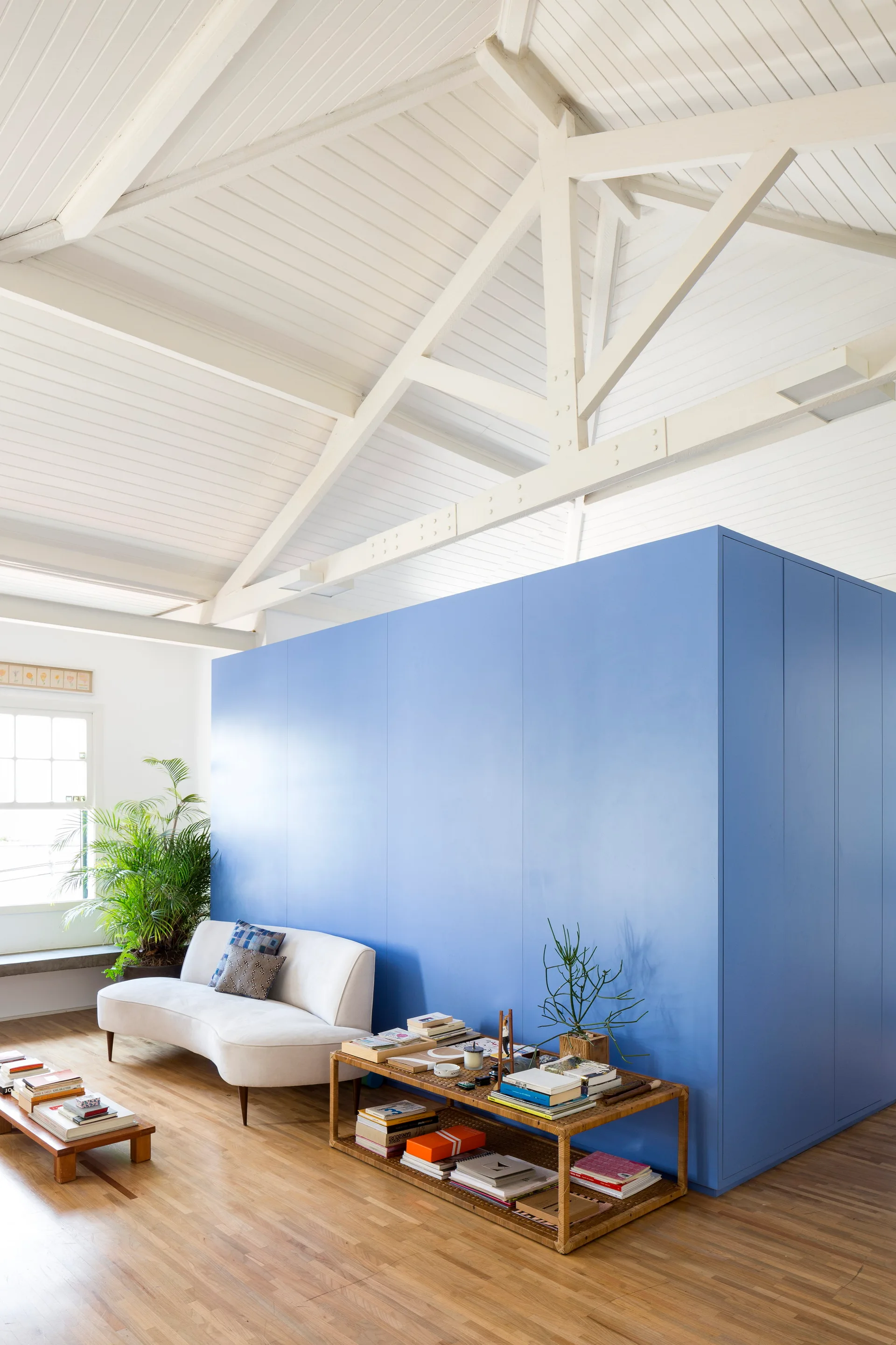 A bright living area features a large blue partition wall, a curved white sofa, a wicker console table with books, and white vaulted ceiling beams.